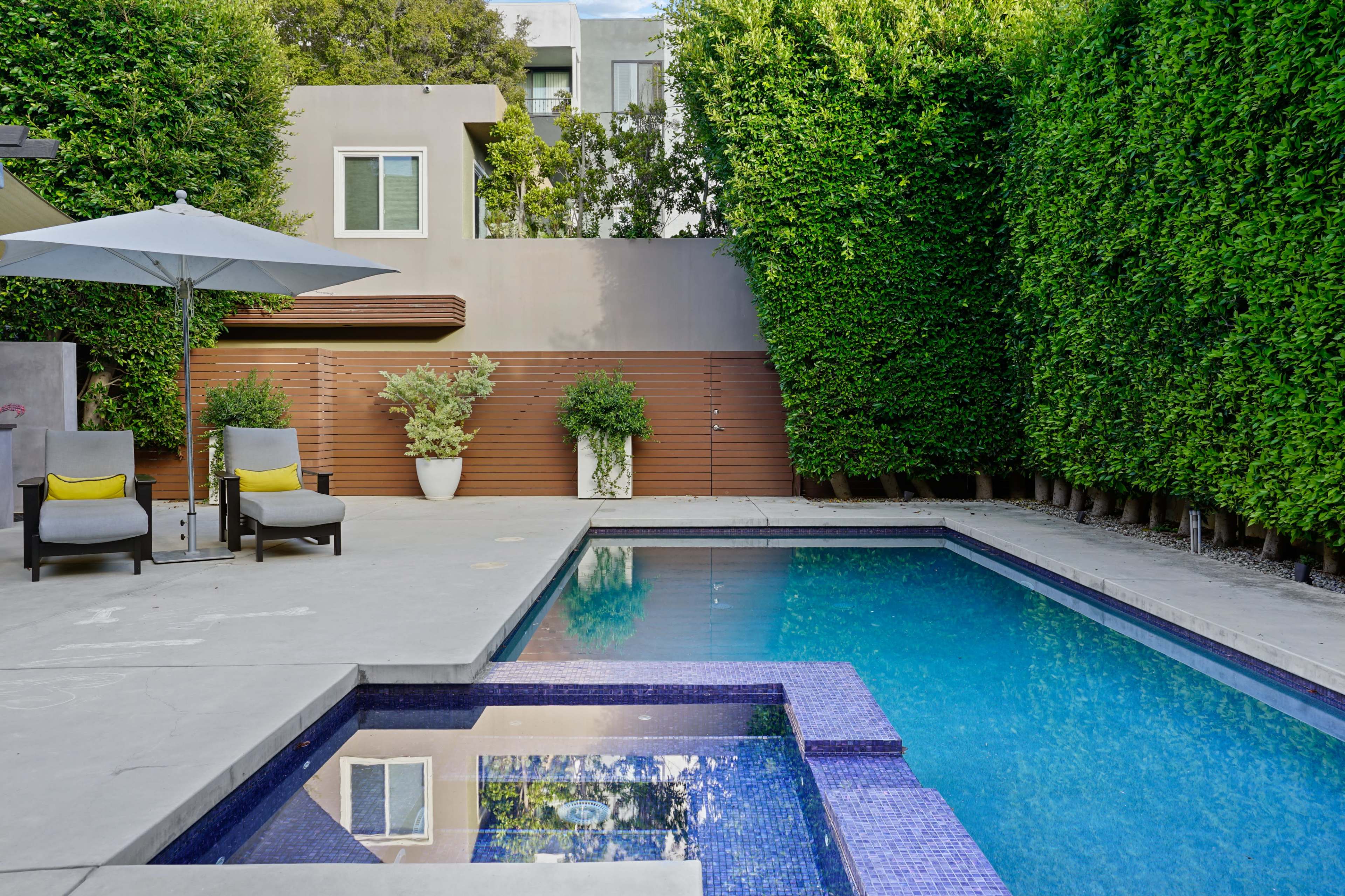 The image shows a modern pool surrounded by neatly trimmed hedges and patio furniture, with a spa built into one corner.
