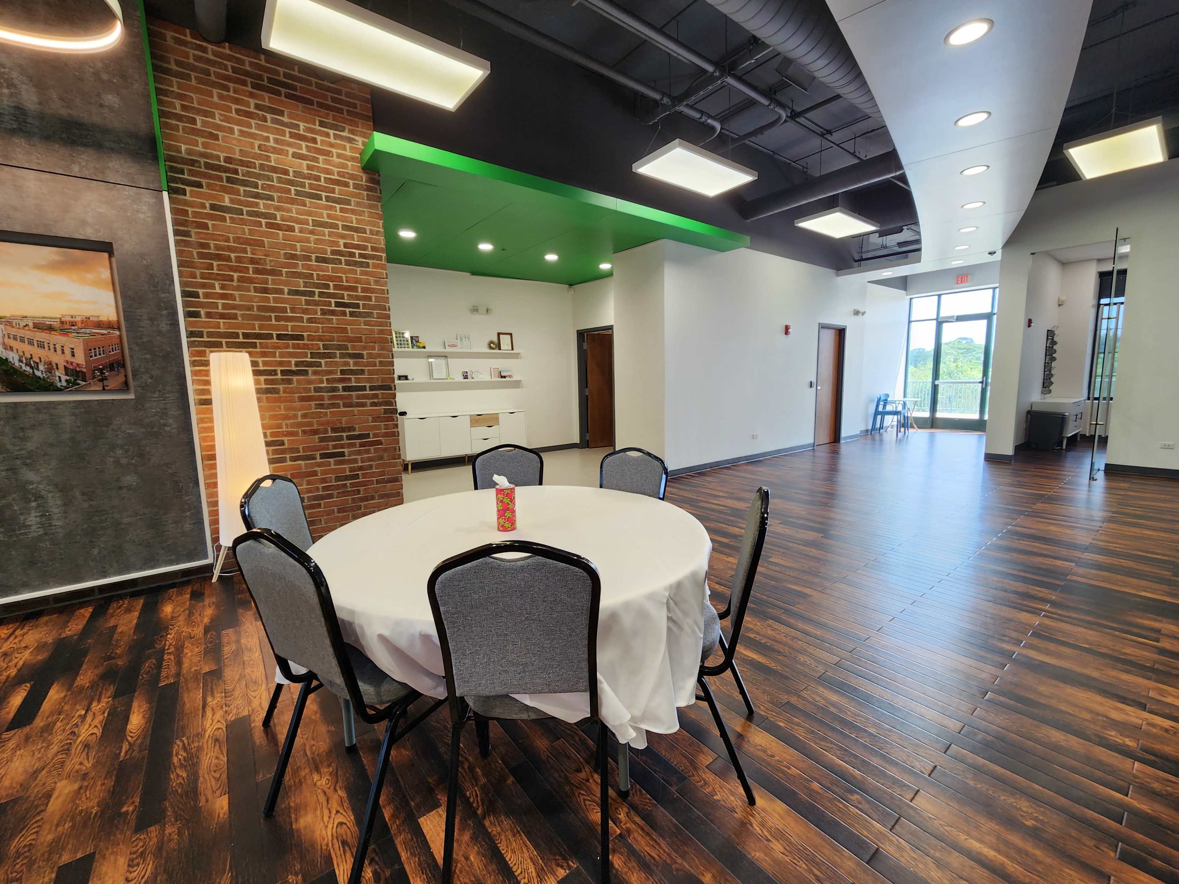 A modern meeting room featuring a round table with a white tablecloth, surrounded by chairs, adjacent to a bright open area with large windows.