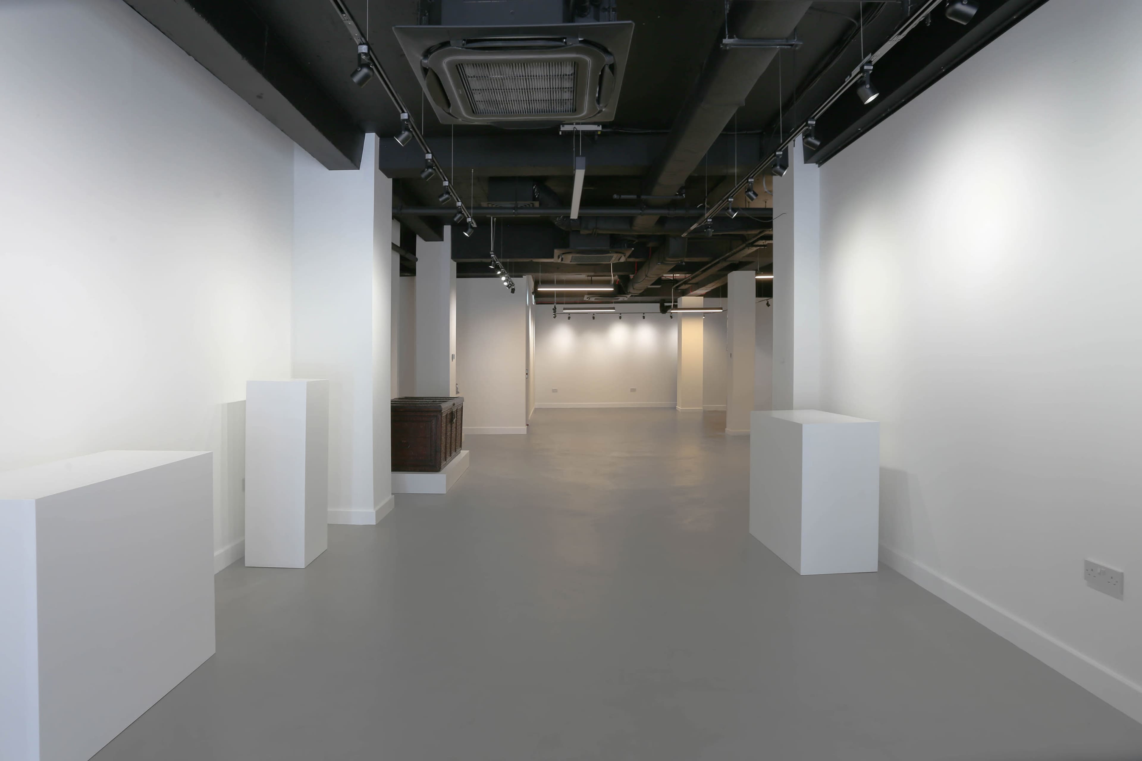 The image shows a spacious, modern interior of an exhibition gallery with white walls, gray flooring, and several white display pedestals.