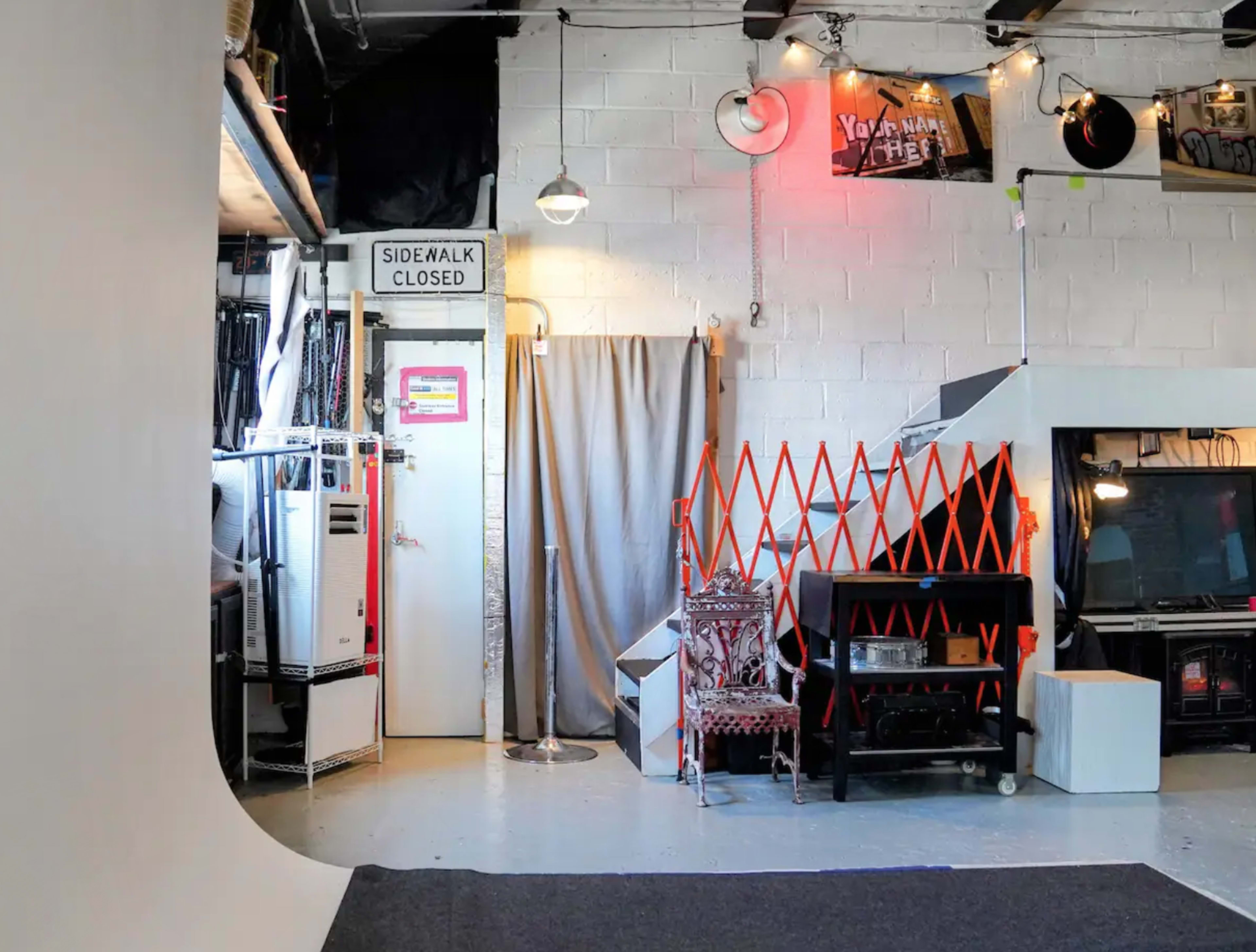 The image shows a studio space with a white backdrop, a closed door labeled "SIDEWALK CLOSED," a curtain, various pieces of furniture, and an orange safety barrier.
