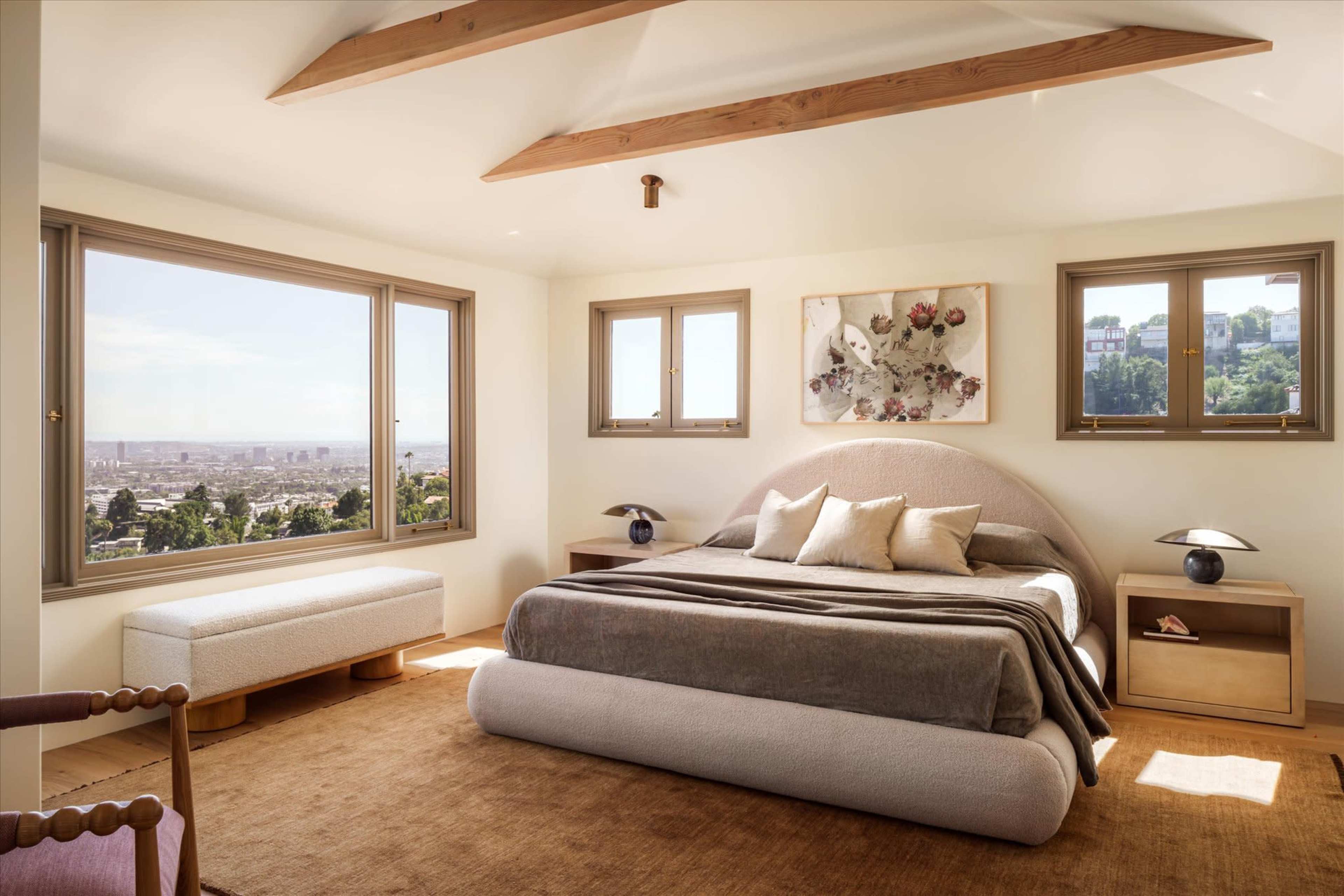 The image shows a modern bedroom with a large bed, a seating area, and windows offering a view of the city.