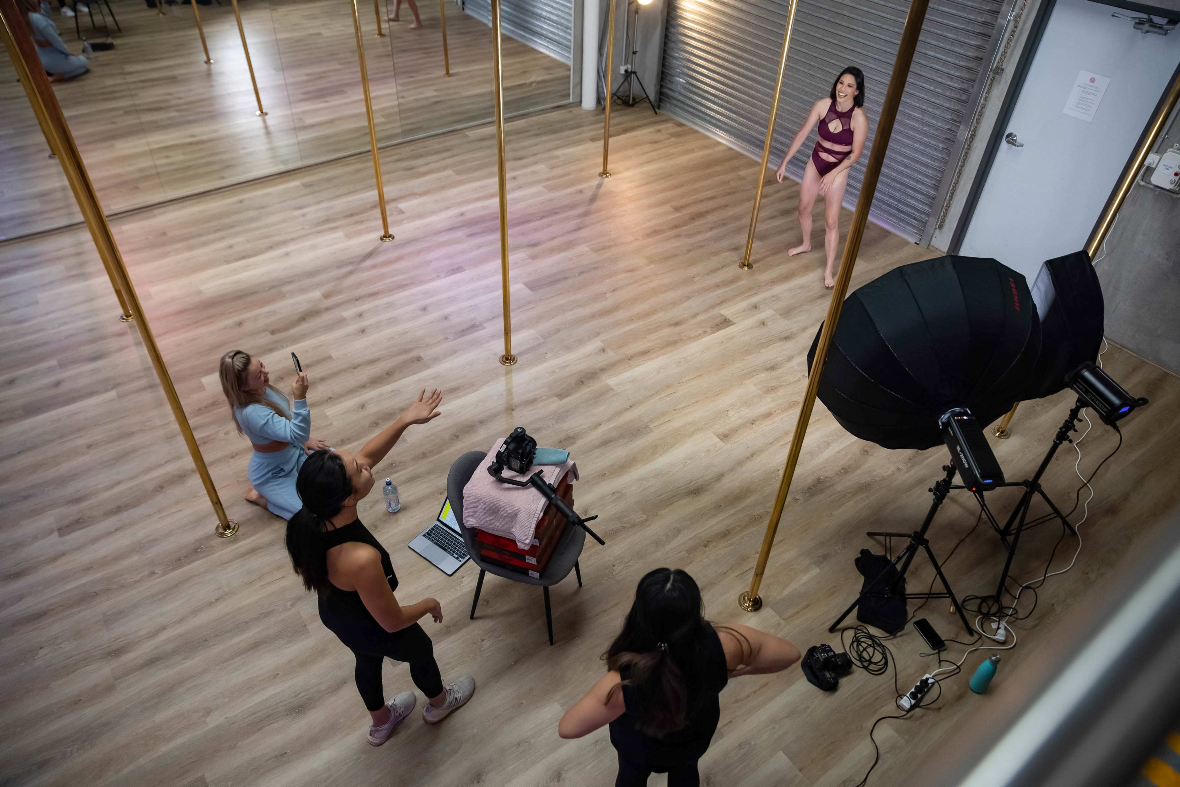 A studio with four women, two standing near a pole while one is sitting behind a camera, and another is gesturing from the side.