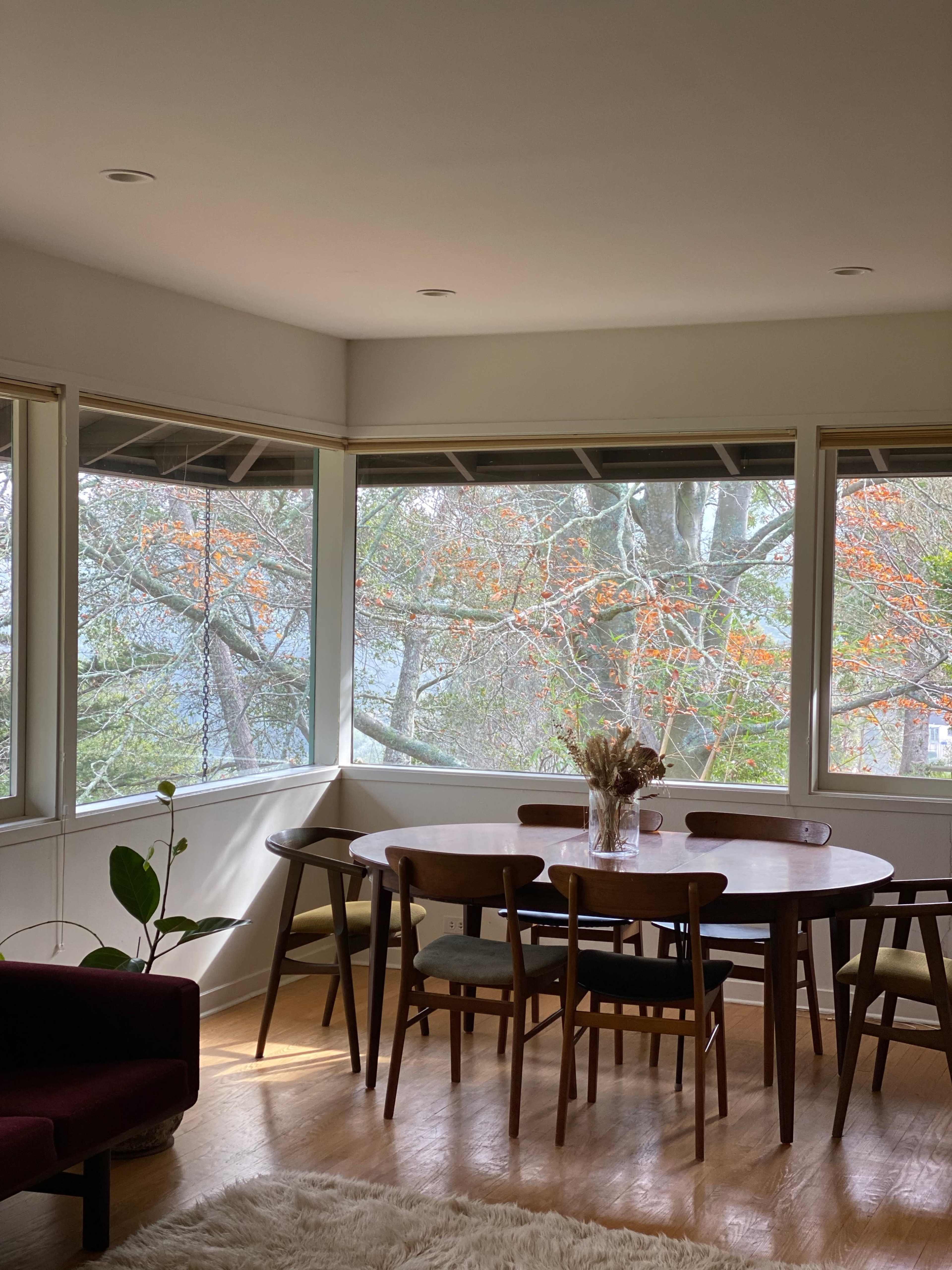 A dining area features a round table surrounded by wooden chairs, with large windows offering a view of trees outside.
