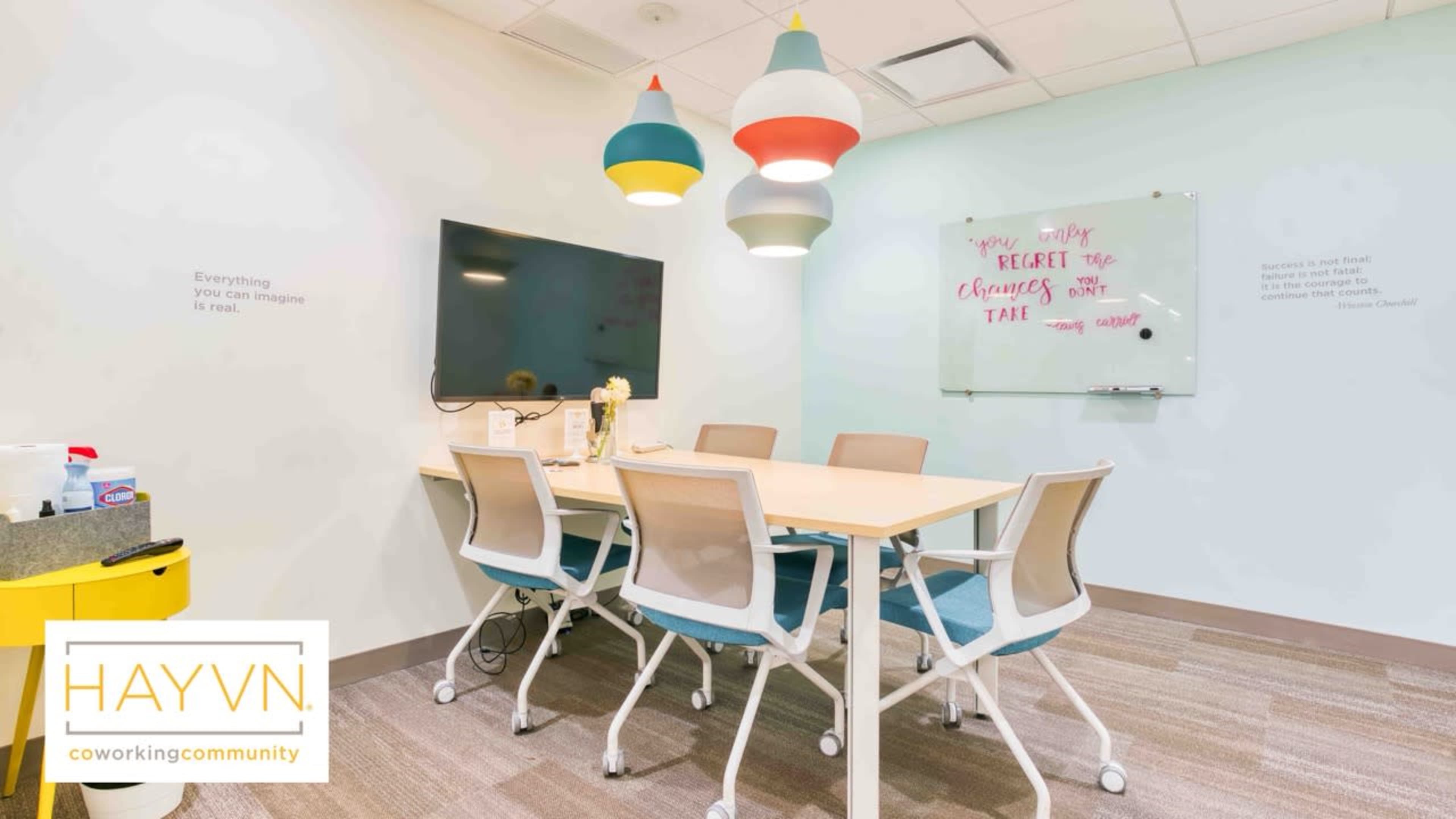 A brightly lit meeting room features a large table surrounded by several chairs, with a television on the wall and colorful pendant lights above.