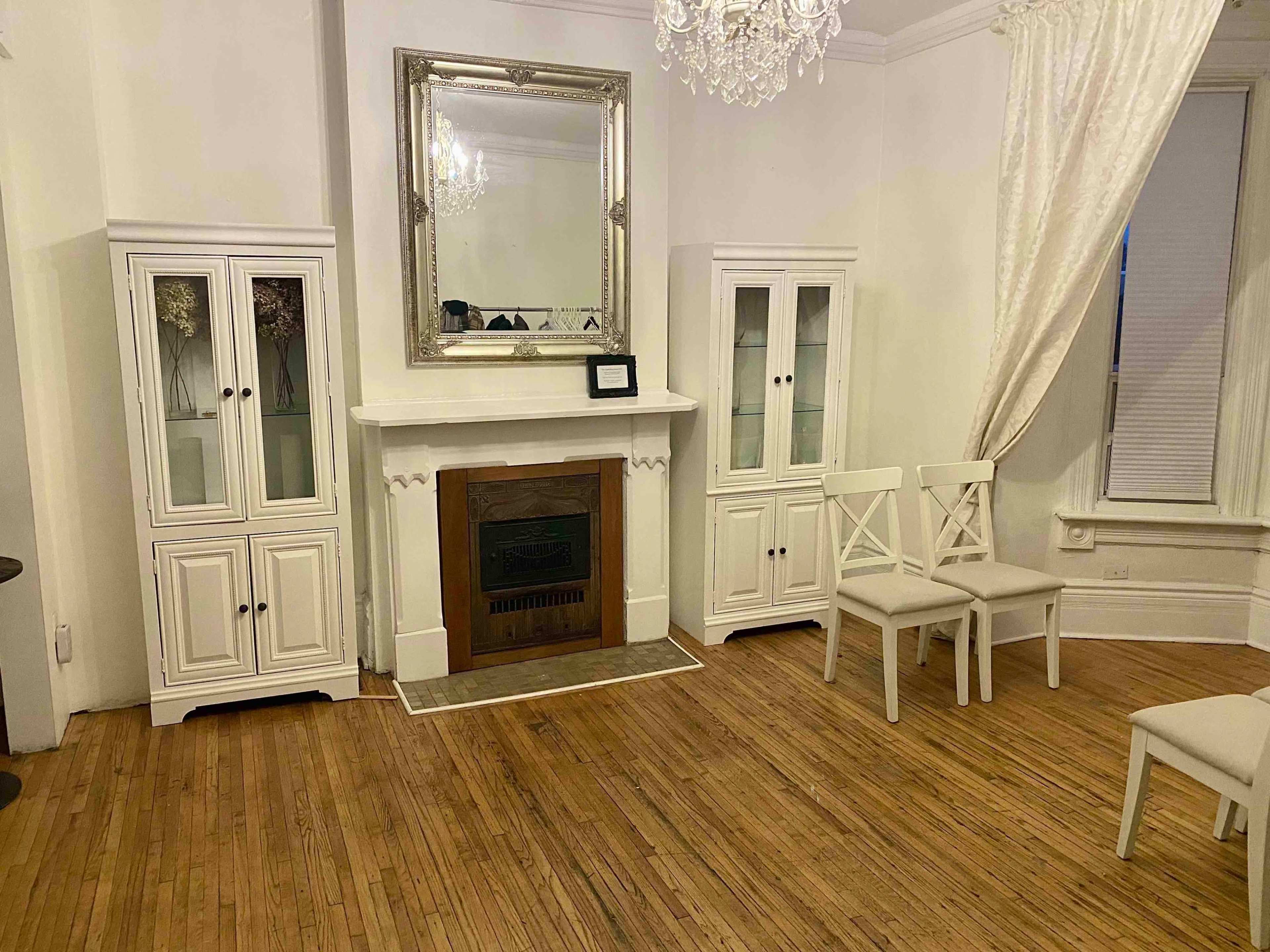 A room with hardwood floors, two white display cabinets flanking a fireplace, and three white chairs positioned nearby.