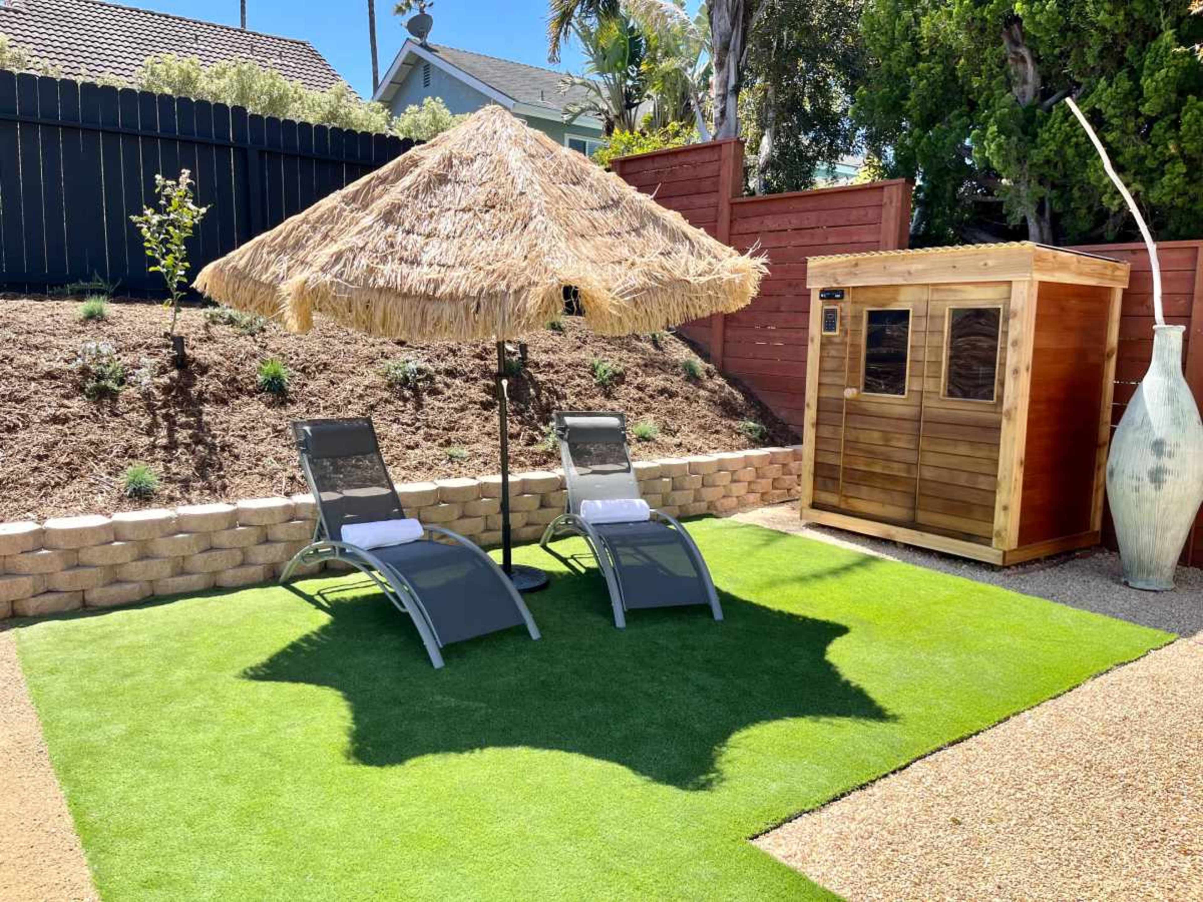 A backyard scene features two lounge chairs under a thatched umbrella on artificial grass, with a wooden shed and decorative vase nearby.