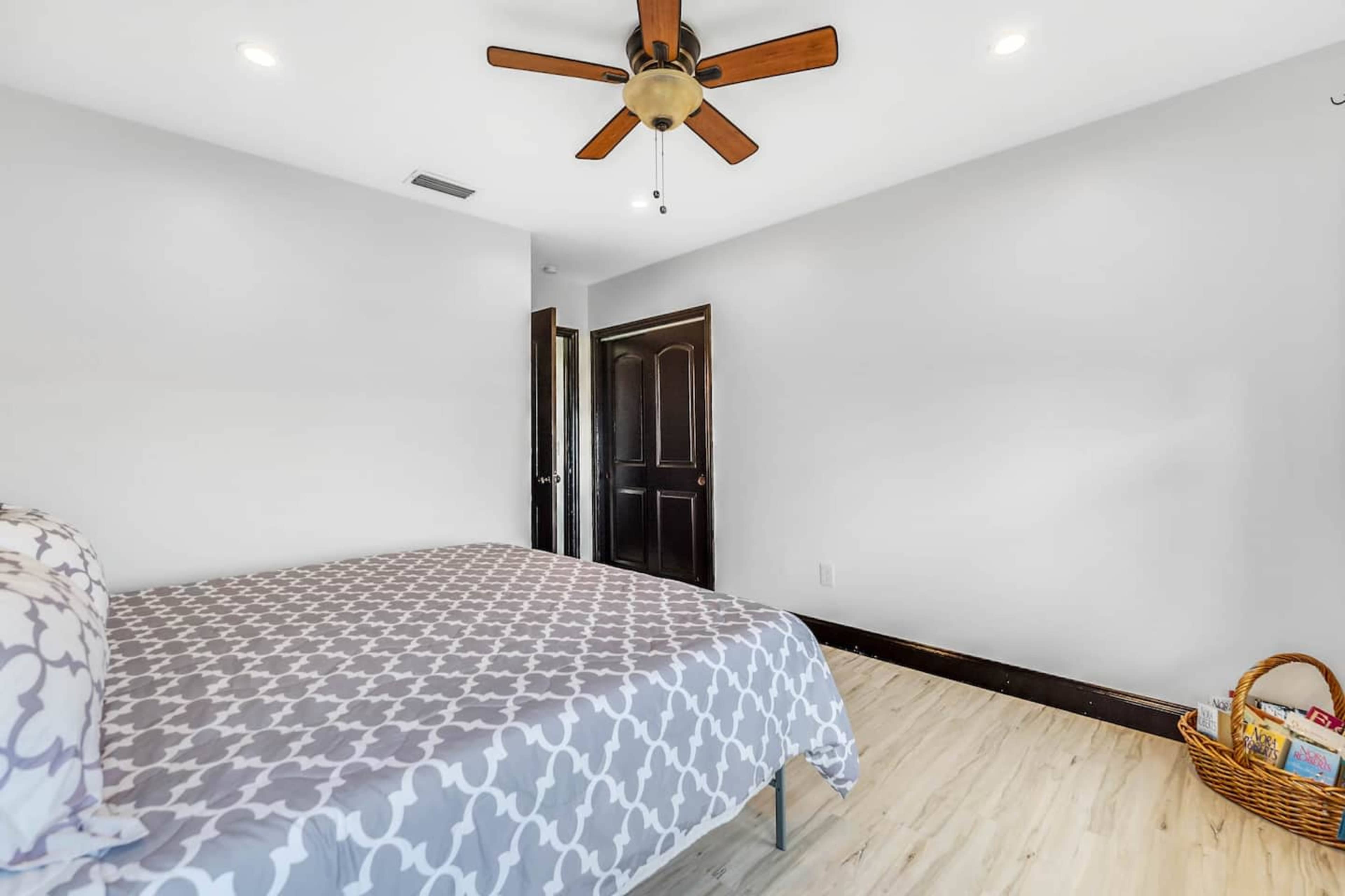 A bedroom features a bed with a patterned duvet, a ceiling fan, and a corner basket filled with books.