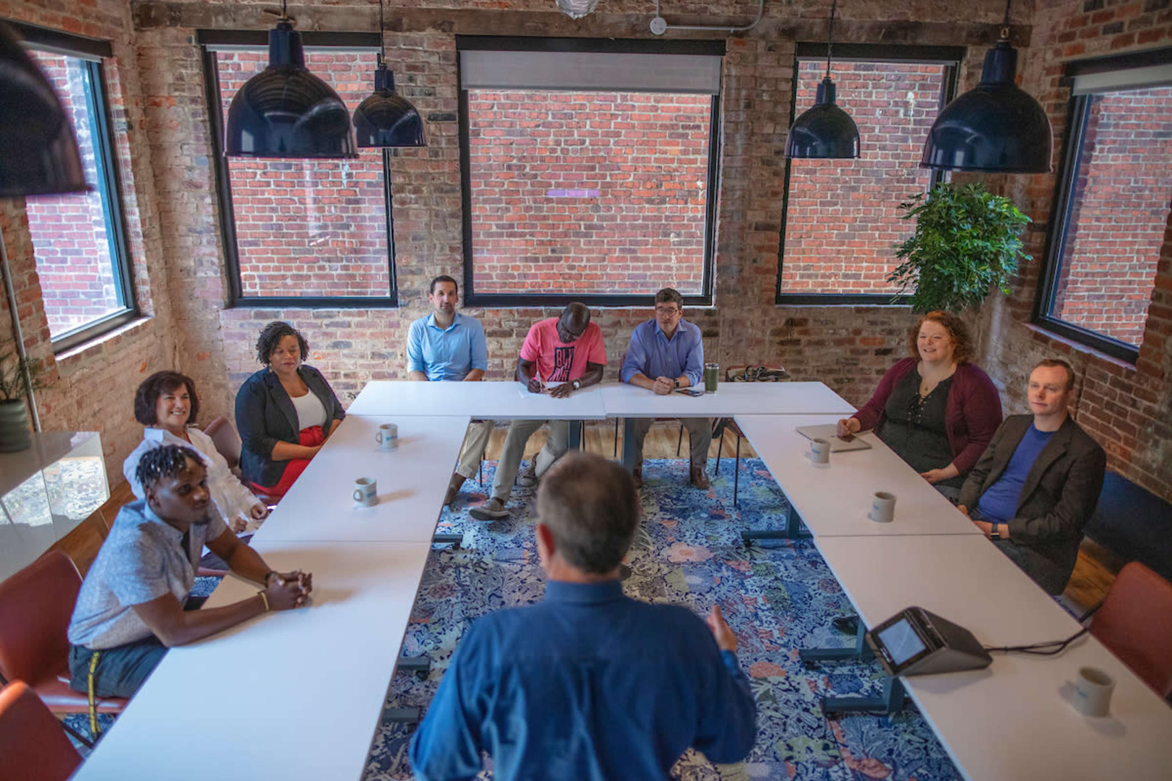 A group of eight people sit around a long table in a brightly lit meeting room with exposed brick walls, while a speaker stands at the front.