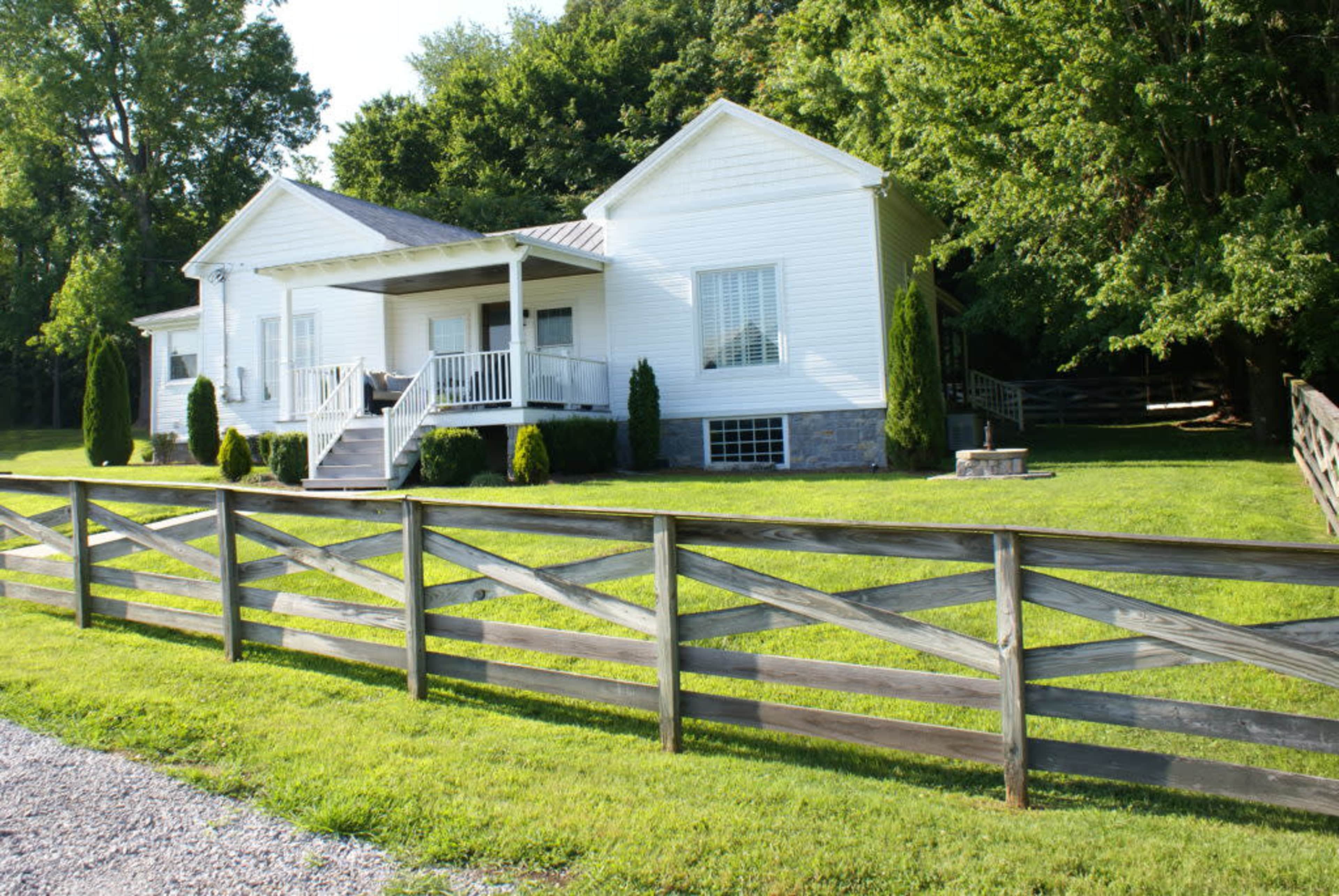 A single-story white house with a front porch is surrounded by a green lawn and a wooden fence.