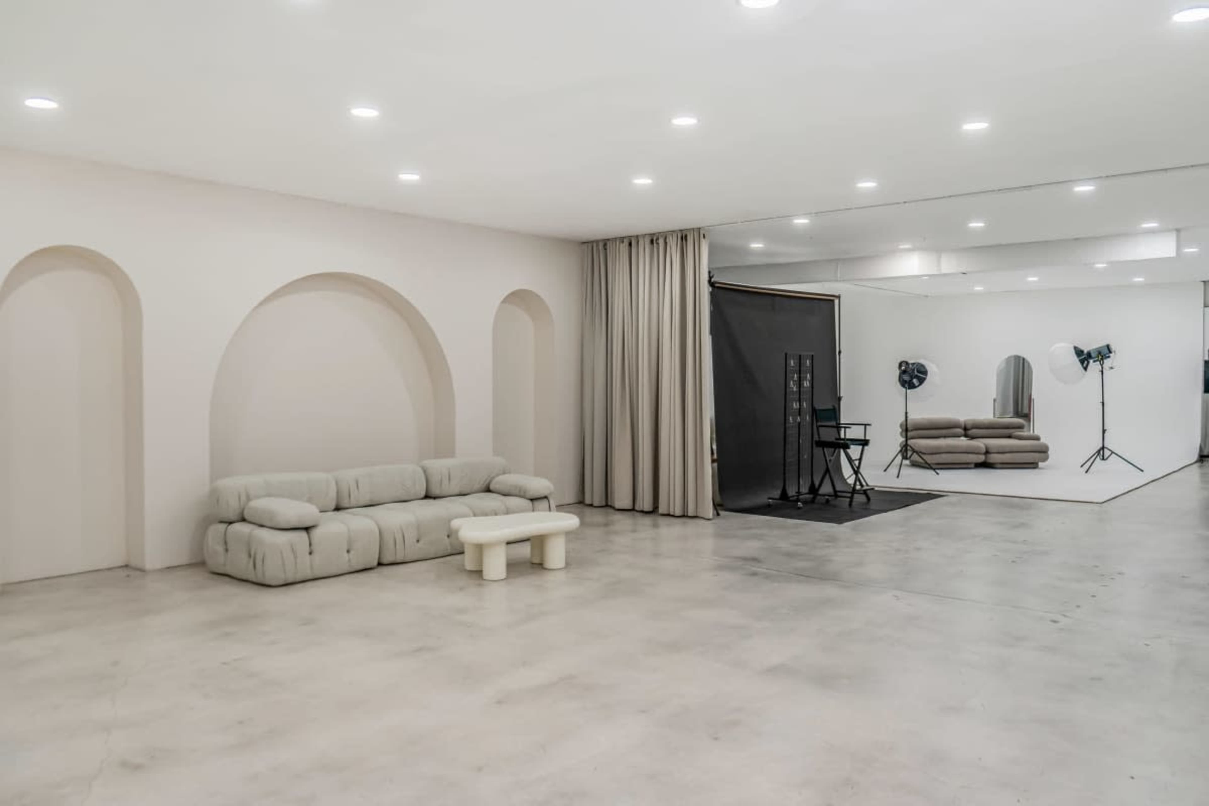 The image shows a minimalist studio space with a light-colored concrete floor, a curved sofa, and a backdrop with lighting equipment set up for photography.
