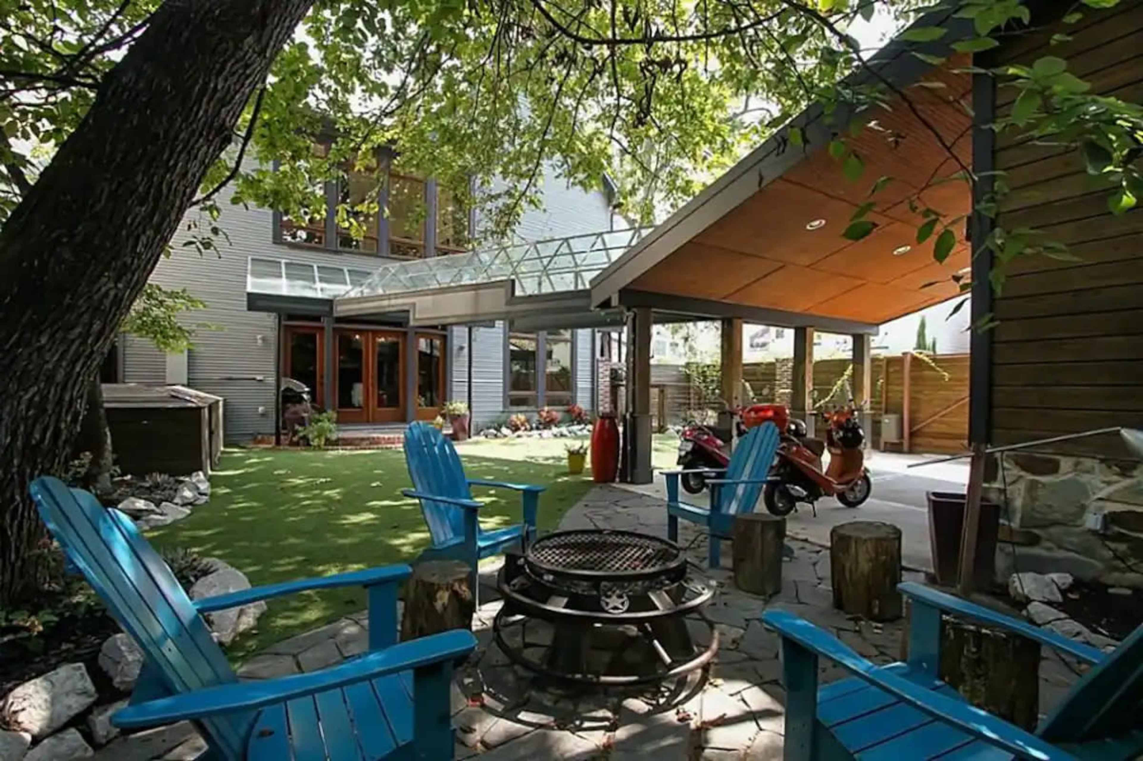 A landscaped backyard with a stone fire pit surrounded by blue chairs and a covered seating area, framed by trees and buildings.
