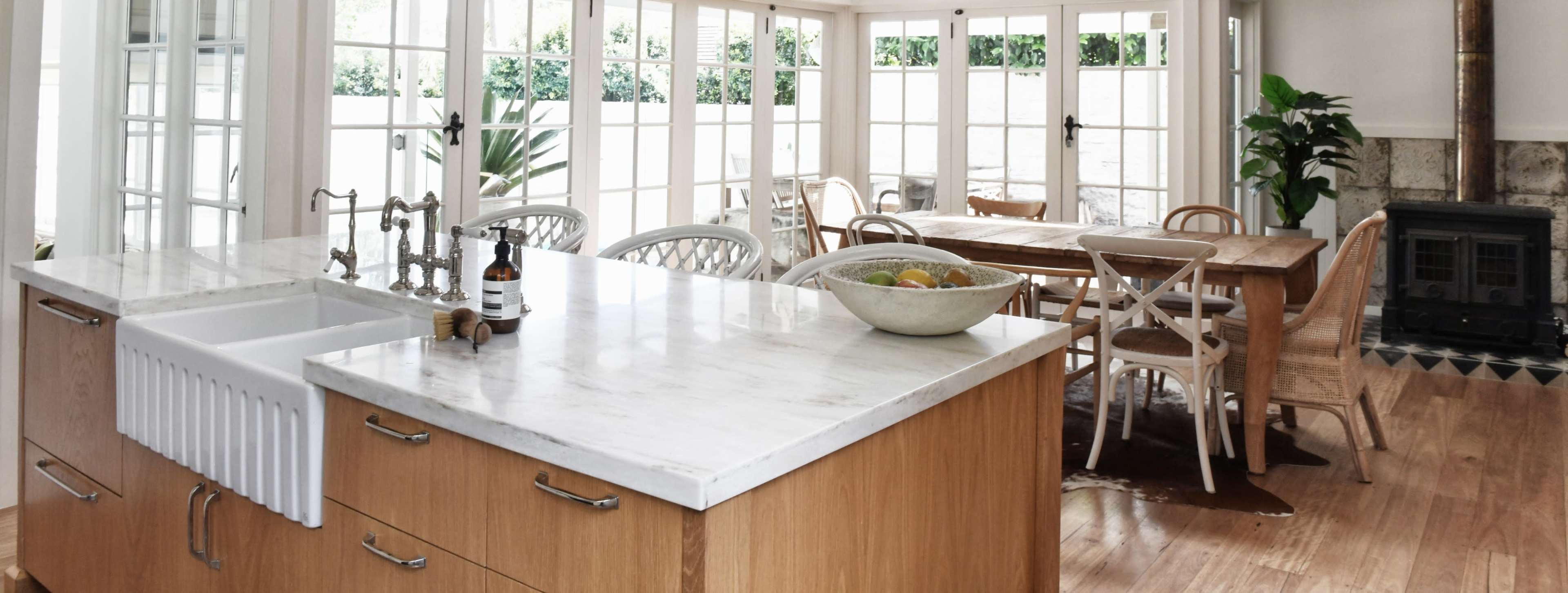 The image shows a modern kitchen with a marble countertop island and a dining area featuring a wooden table and chairs, illuminated by large windows.