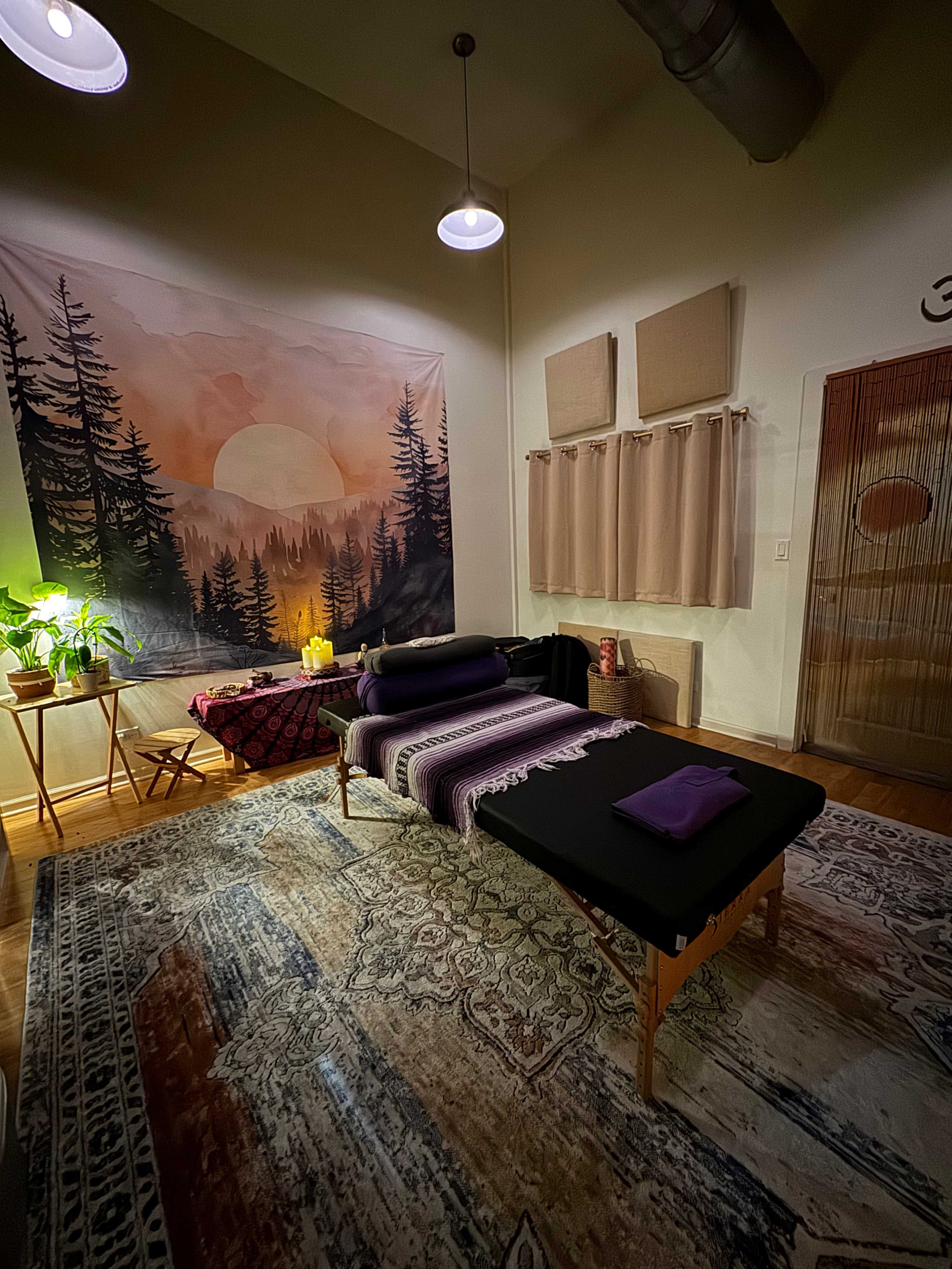 The image shows a serene treatment room featuring a massage table, ambient lighting, a large decorative wall tapestry depicting a sunset, and various plants and accessories.
