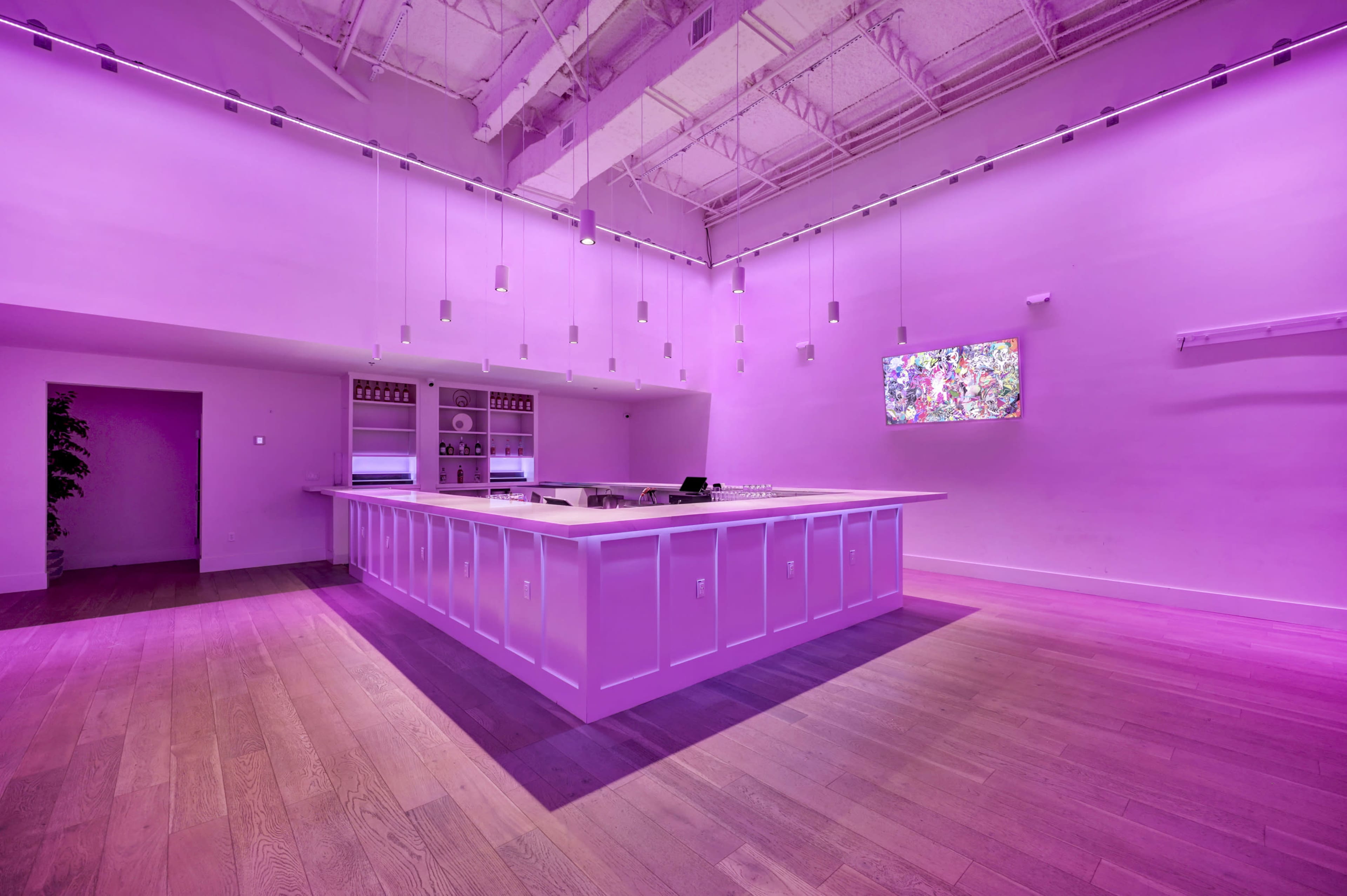 A minimalist kitchen space with a large white island illuminated by purple lighting, featuring a wall-mounted screen and wooden flooring.
