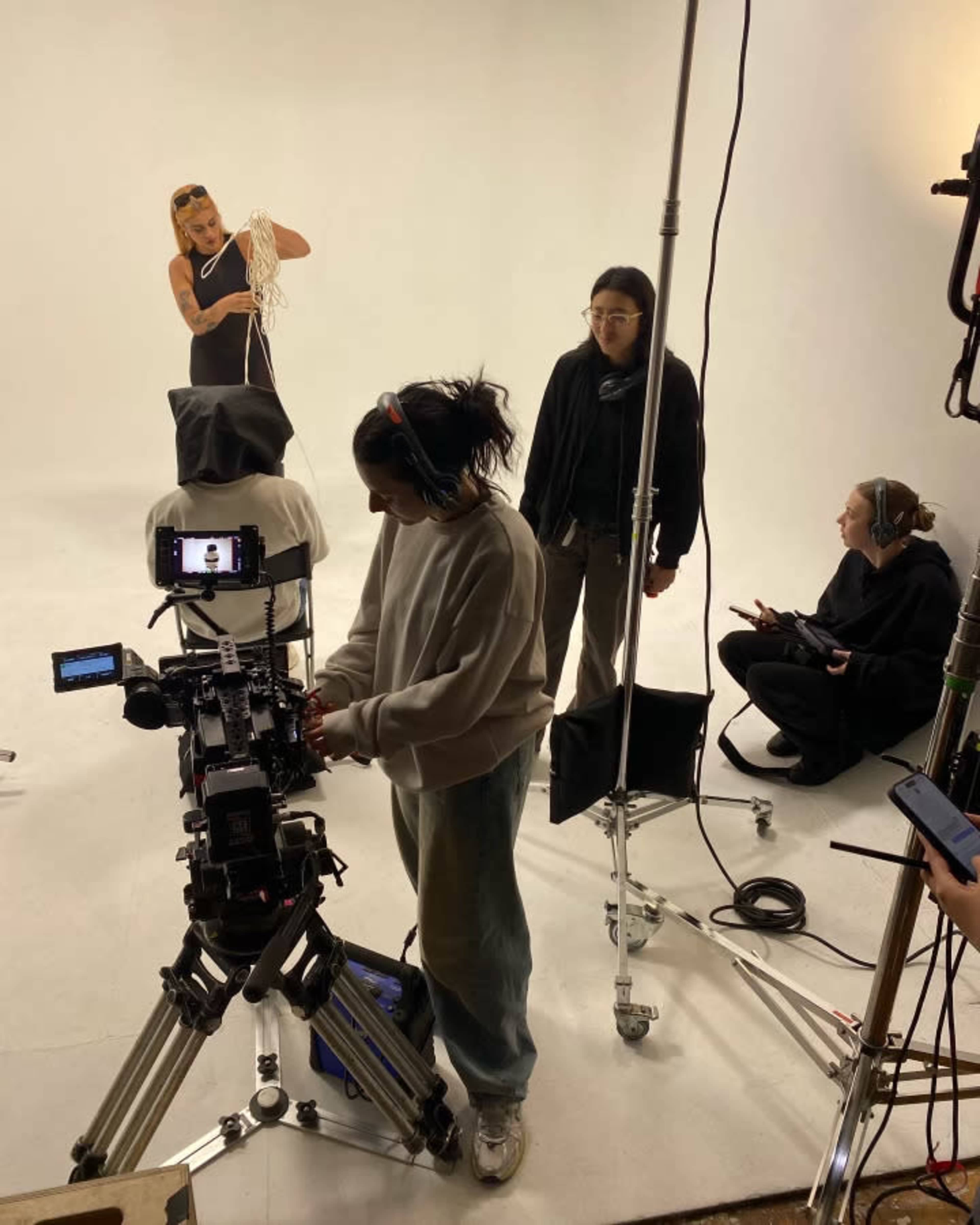 A group of people is filming a production in a studio with a white backdrop, featuring a person performing with a rope while others manage cameras and equipment.