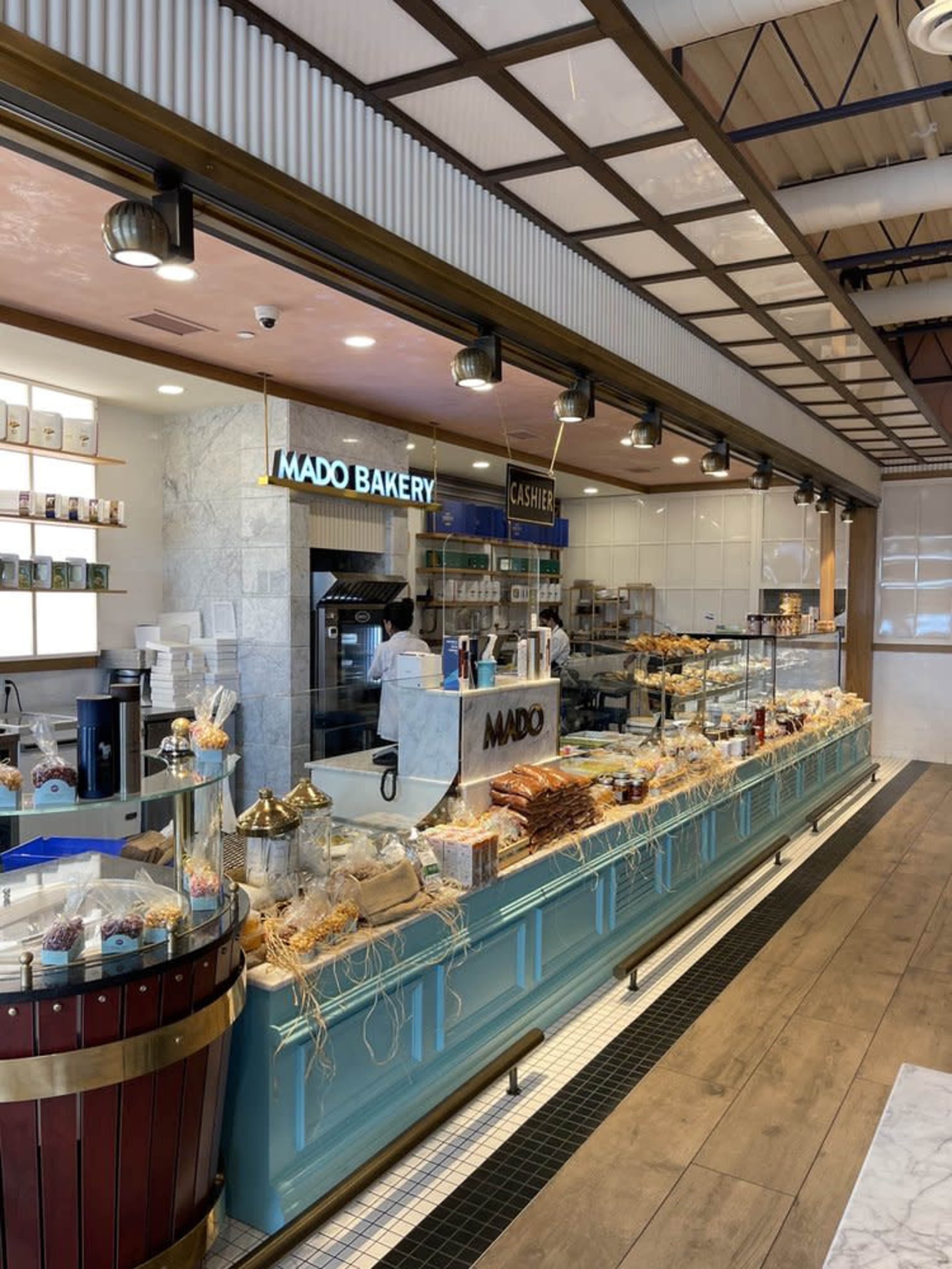 The image shows a brightly lit bakery displaying a variety of baked goods and desserts behind a glass counter, with a cash register area visible in the background.