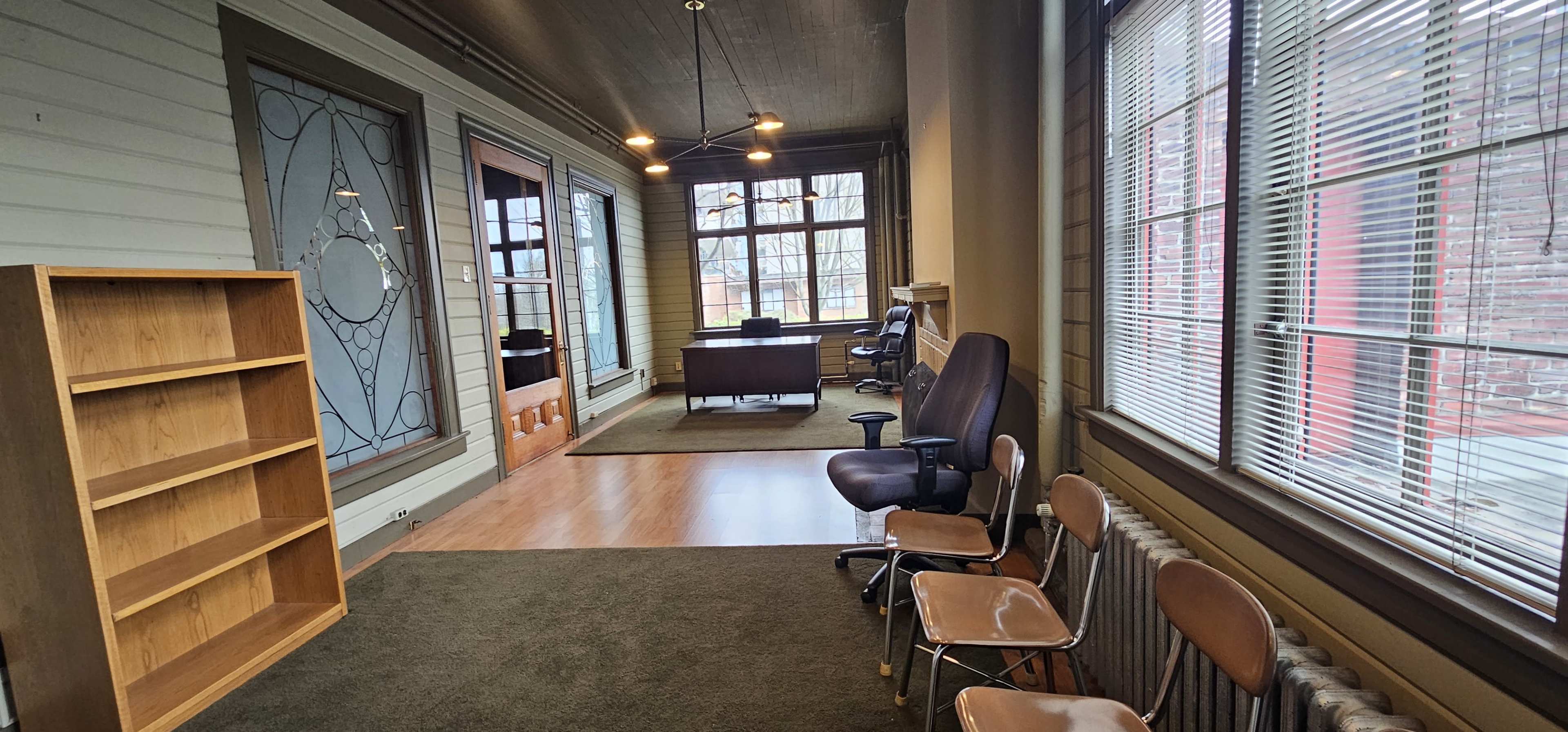 A hallway with a wooden bookshelf, several chairs, and large windows illuminating a desk area in the background.