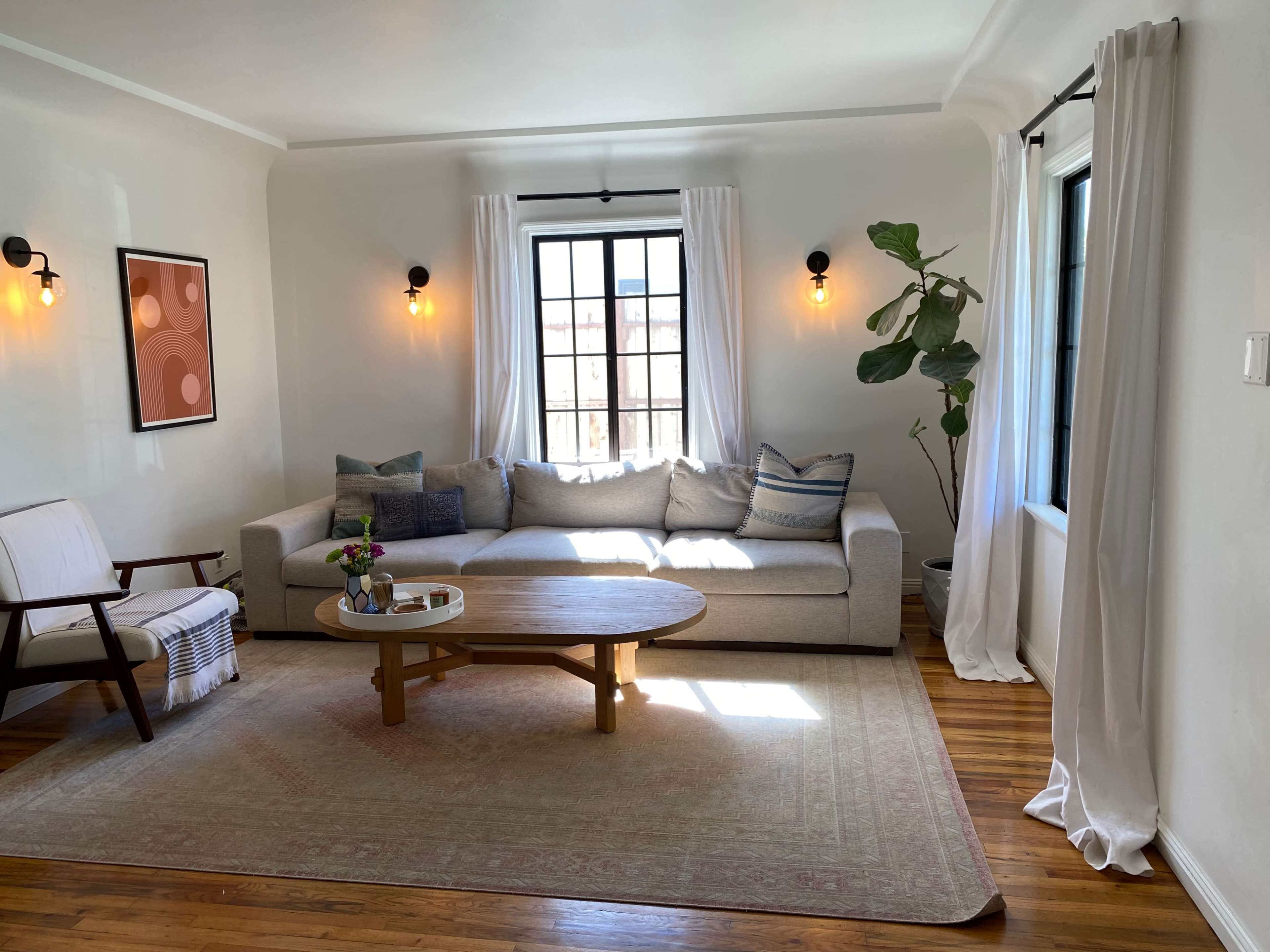 The living room features a light-colored sofa, a wooden coffee table, and two large windows with sheer curtains, complemented by a potted plant and a wall art piece.