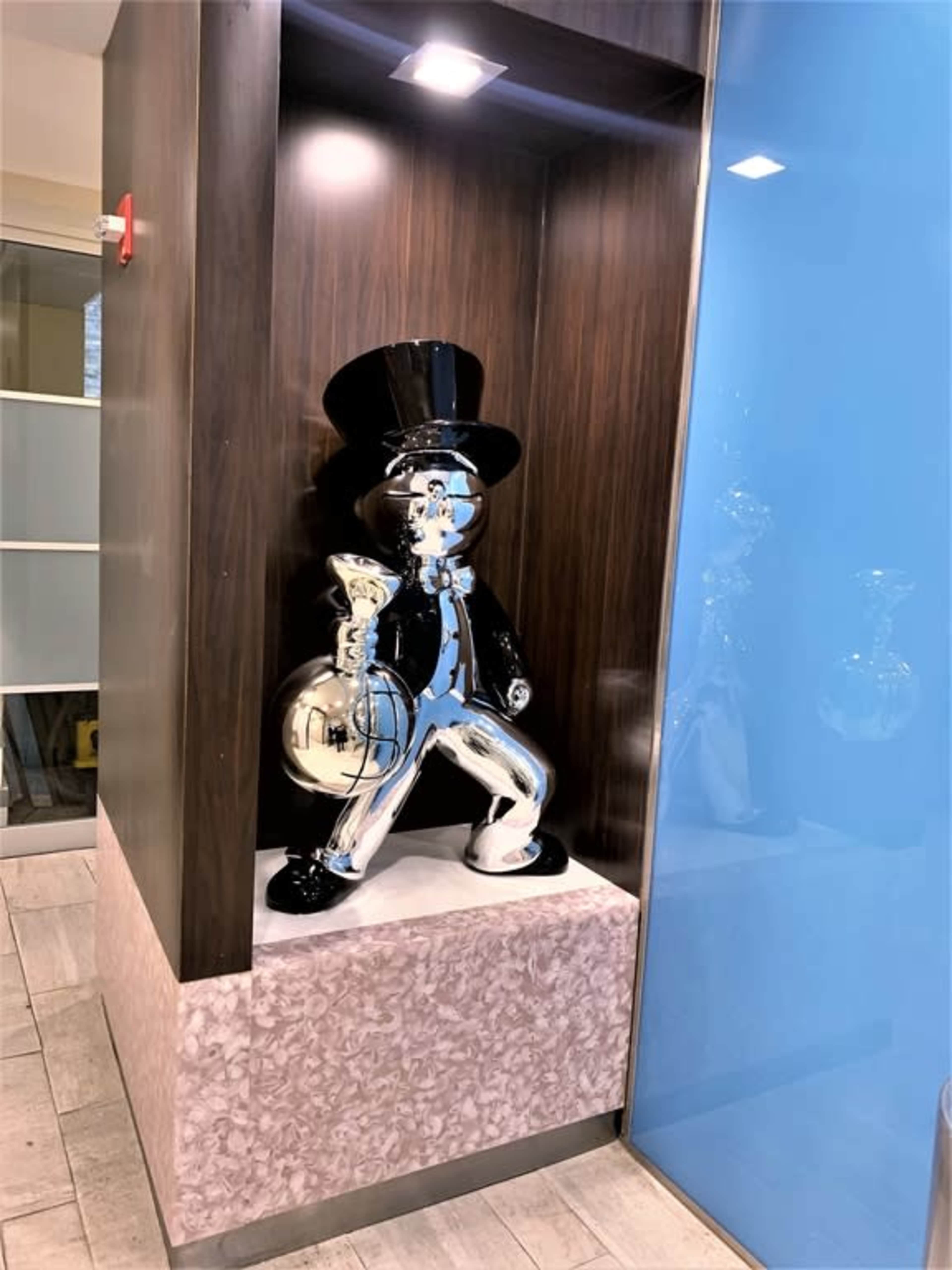 A shiny, metallic sculpture of a top-hatted character holding a bag is displayed in a nook with a contrasting blue wall.
