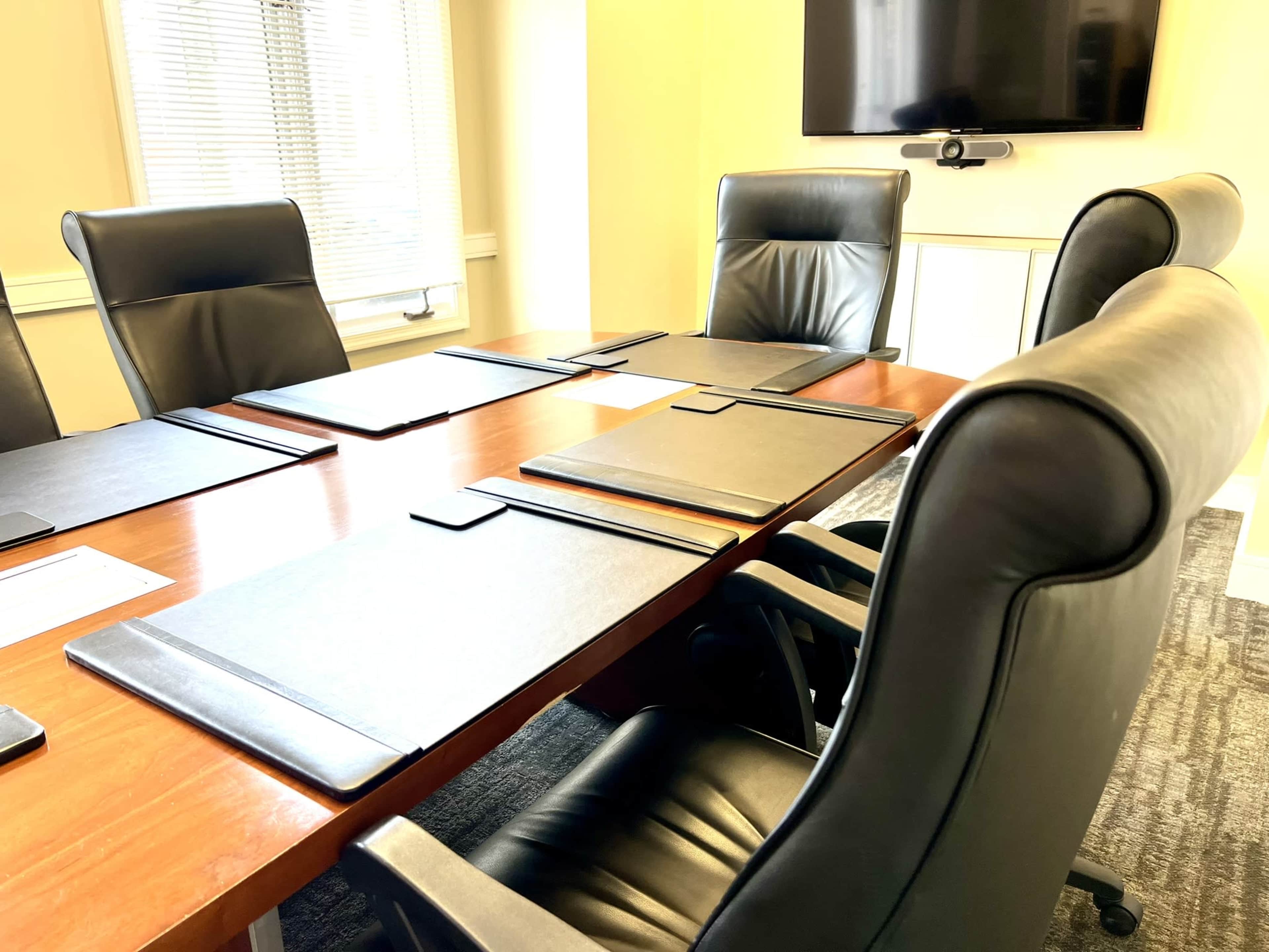 A conference room table with several black folders and four leather chairs is shown, with a television mounted on the wall in the background.