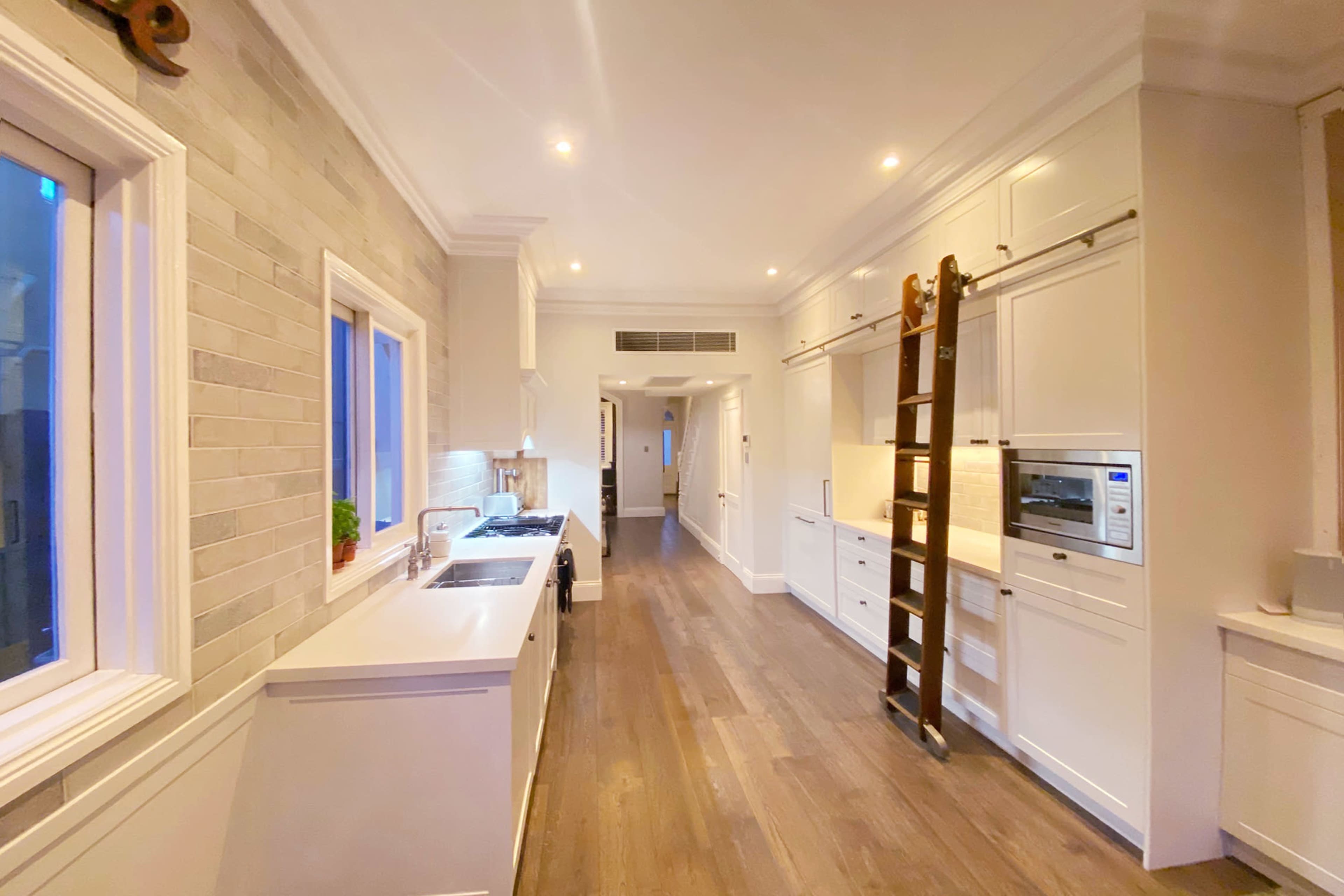 The image shows a modern kitchen with white cabinetry, a large window, and a wooden ladder against the wall.