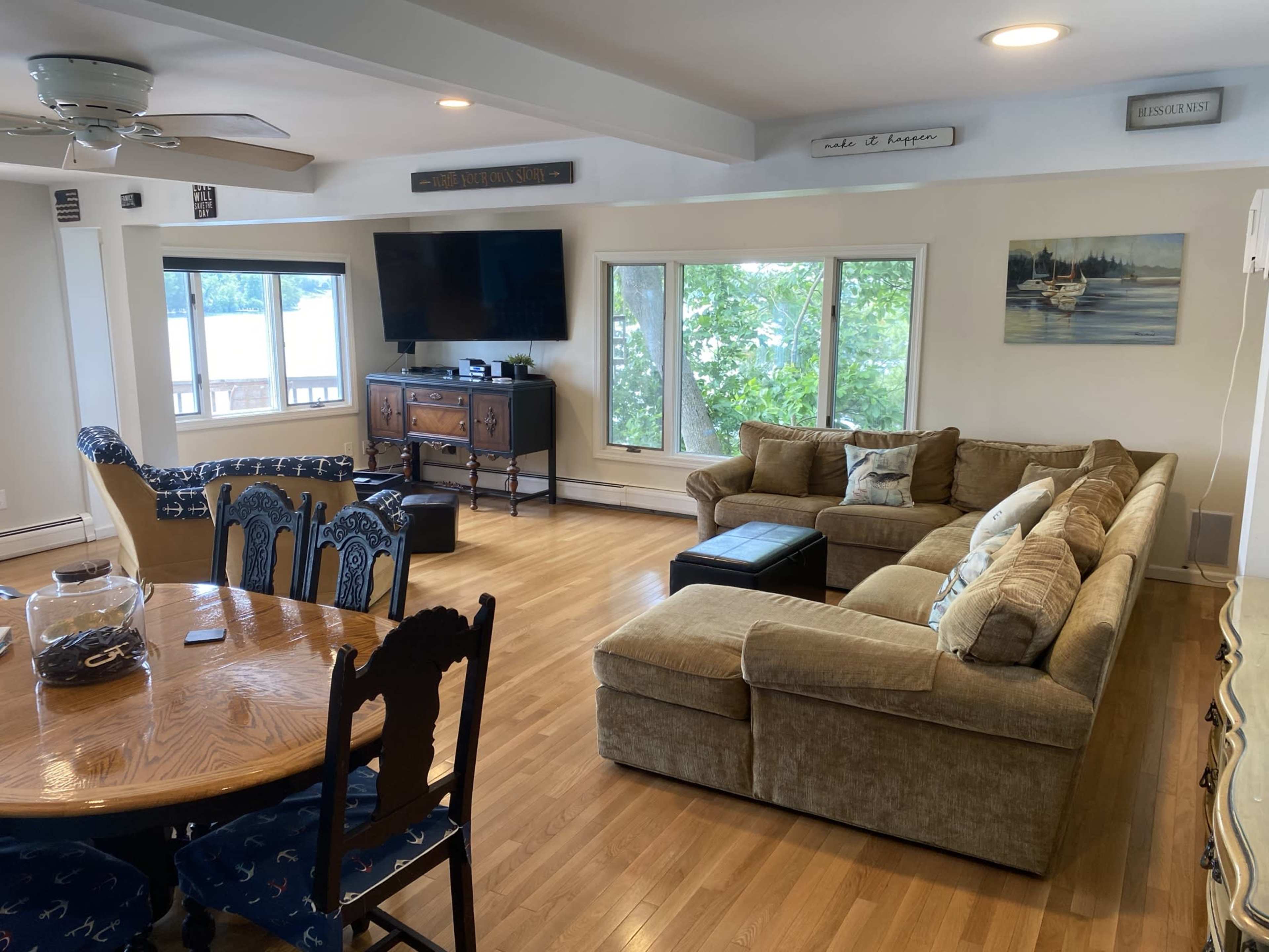 The image shows a spacious living area featuring a sectional sofa, a coffee table, a television, and large windows overlooking a body of water.