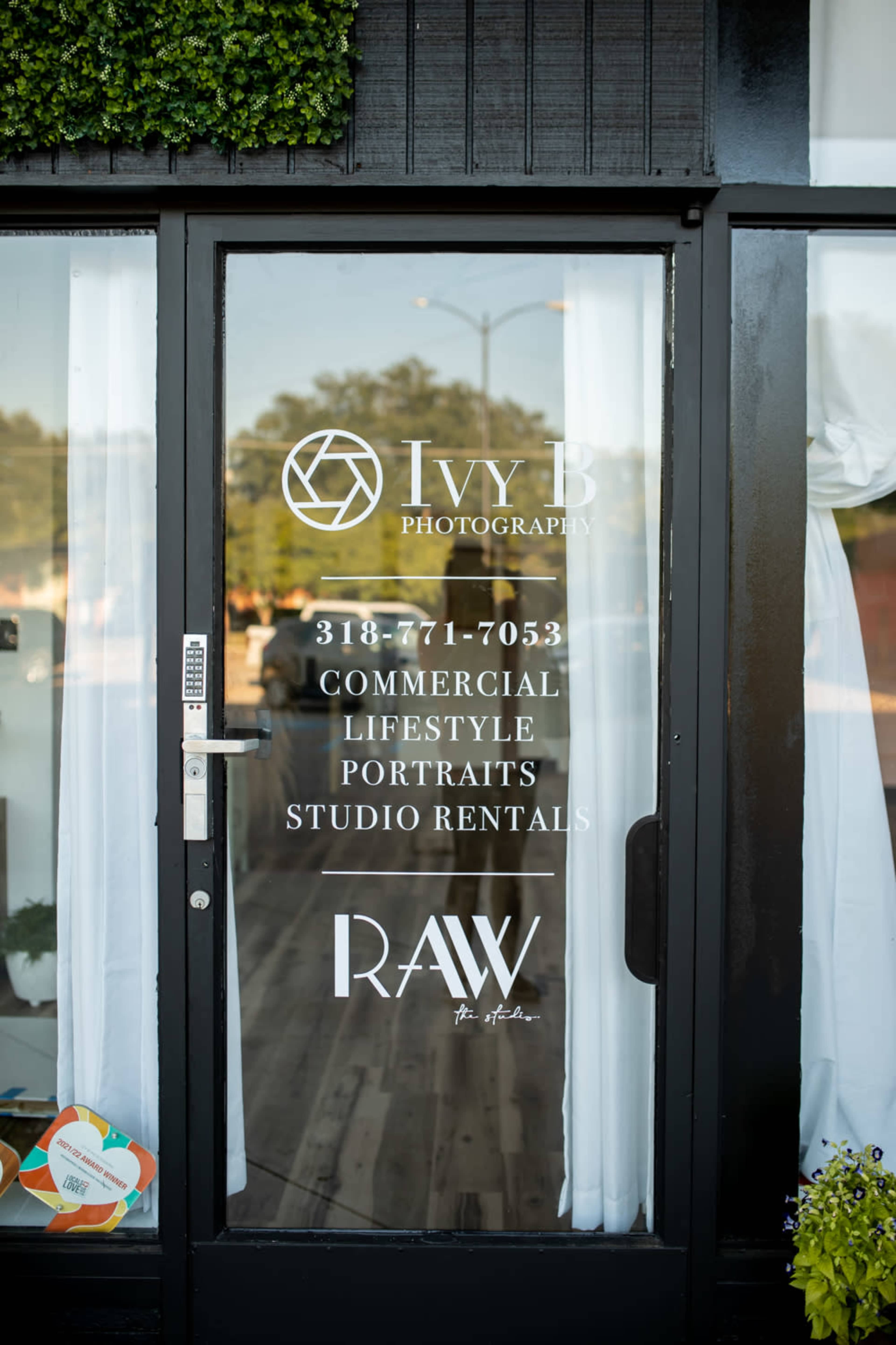 The image shows the glass entrance door of Ivy B Photography, featuring a sign with contact information and services offered, along with the logo of RAW.
