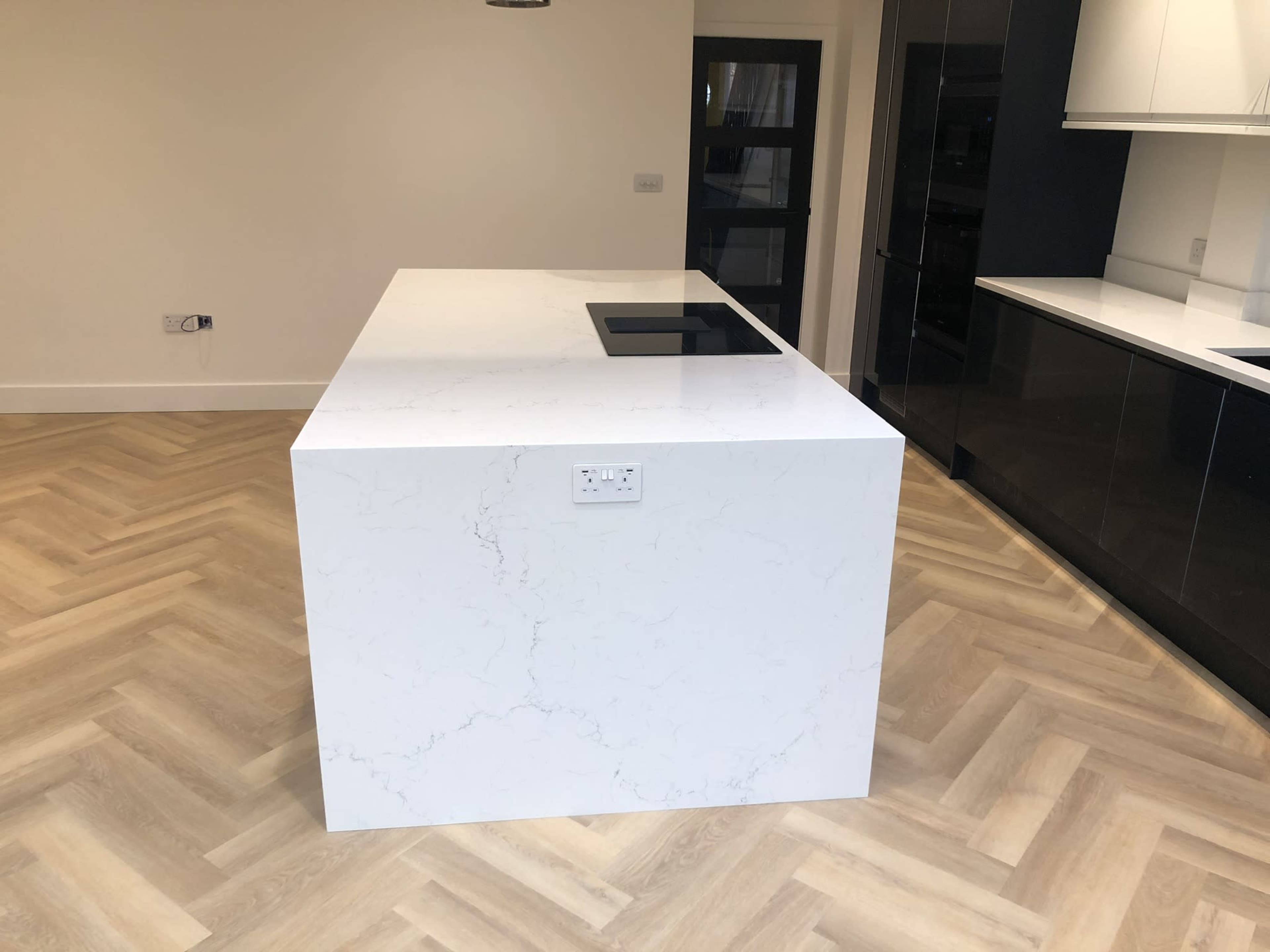 The image shows a modern kitchen with a large white quartz island and a built-in cooktop, set against a herringbone-patterned wooden floor.