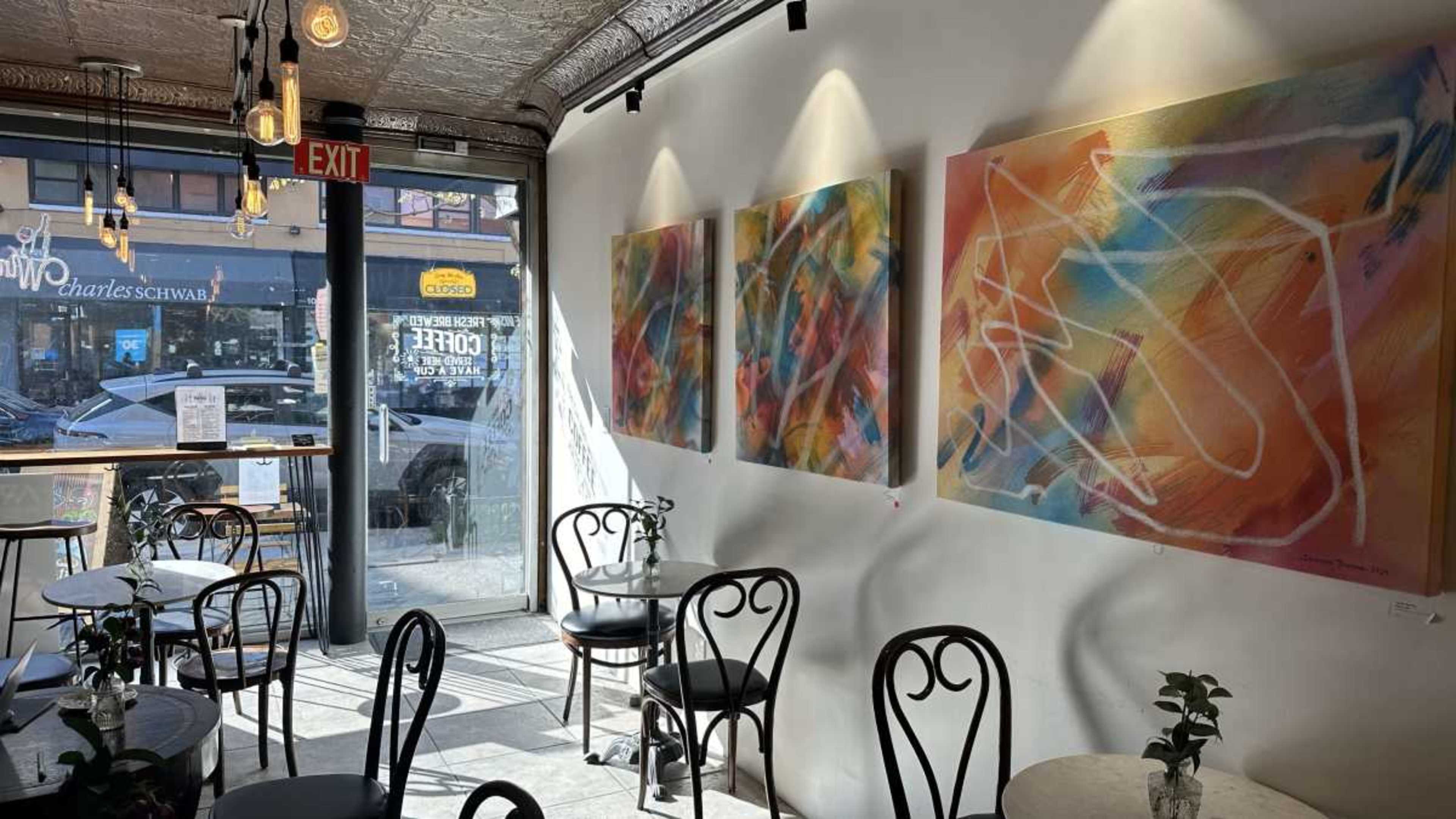 A bright cafe features several abstract paintings on the wall next to a doorway, with tables and chairs arranged throughout the space.
