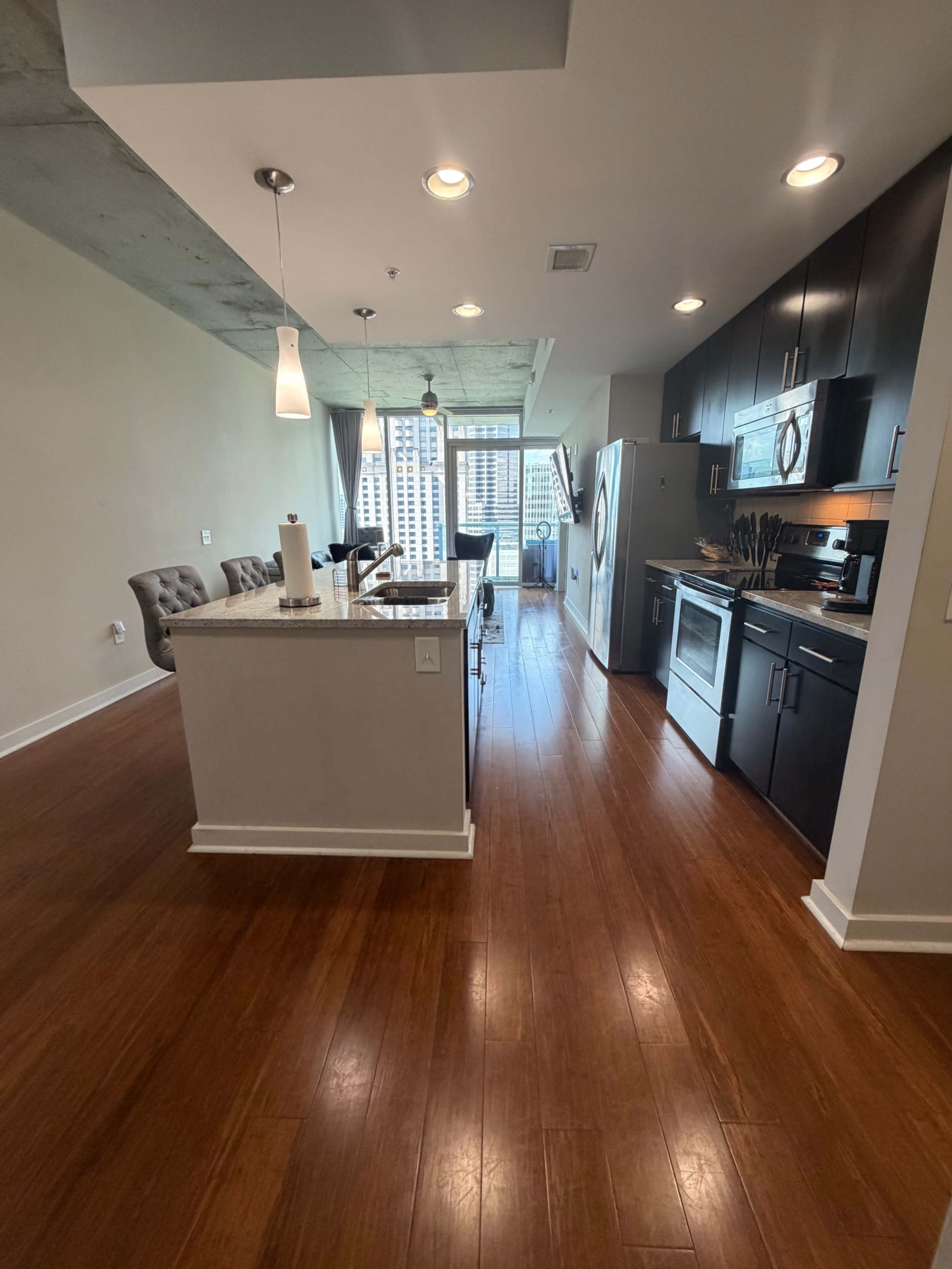 The image shows a modern kitchen and dining area featuring dark cabinetry, stainless steel appliances, and a view through large windows.