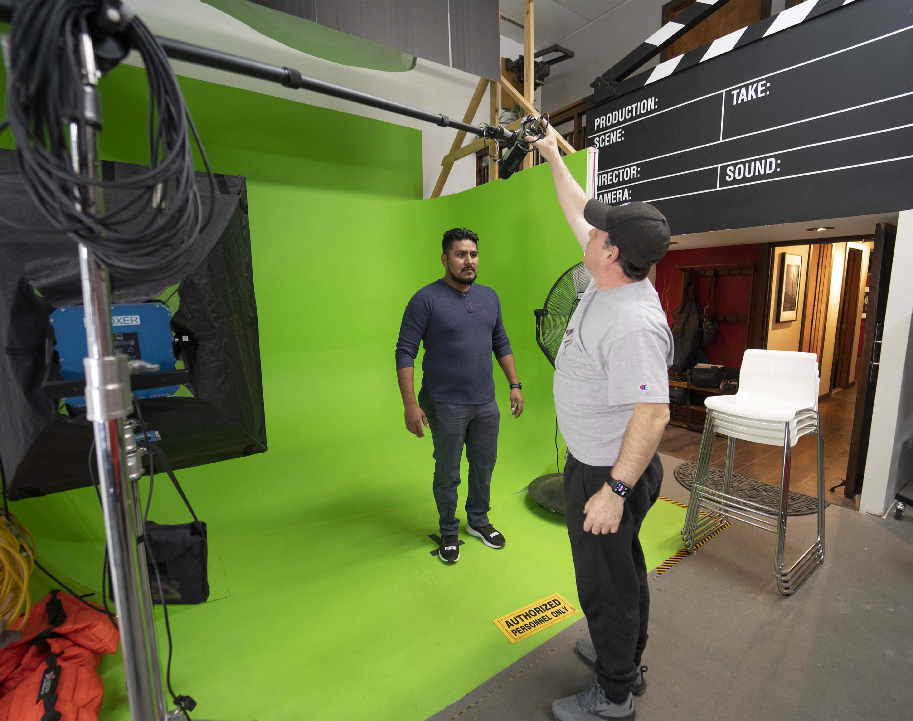 A man stands on a green backdrop in a studio while another man adjusts a camera on a tripod.