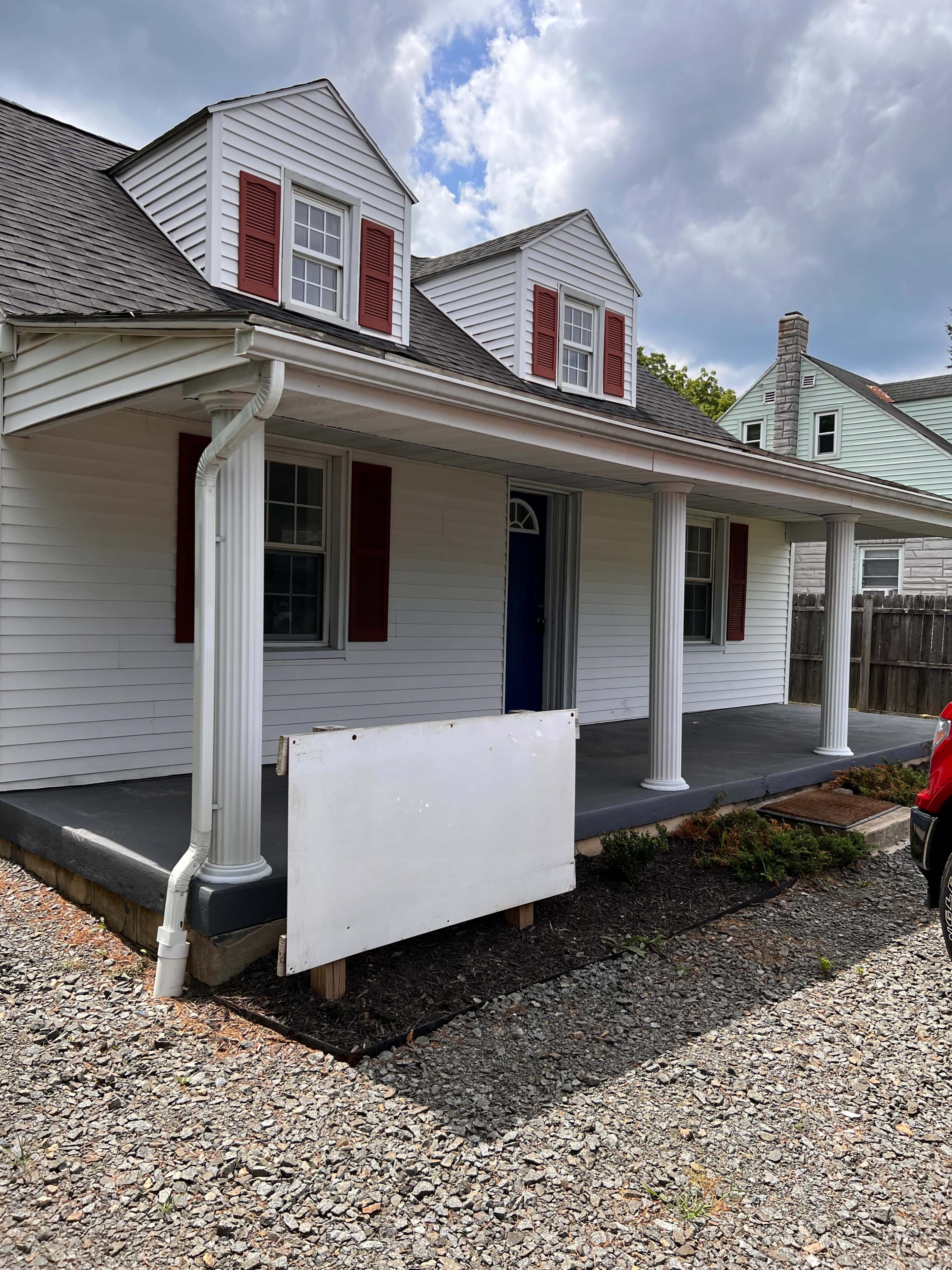 A white house with red shutters, featuring a front porch supported by two columns, and a blank sign leaning against the porch.