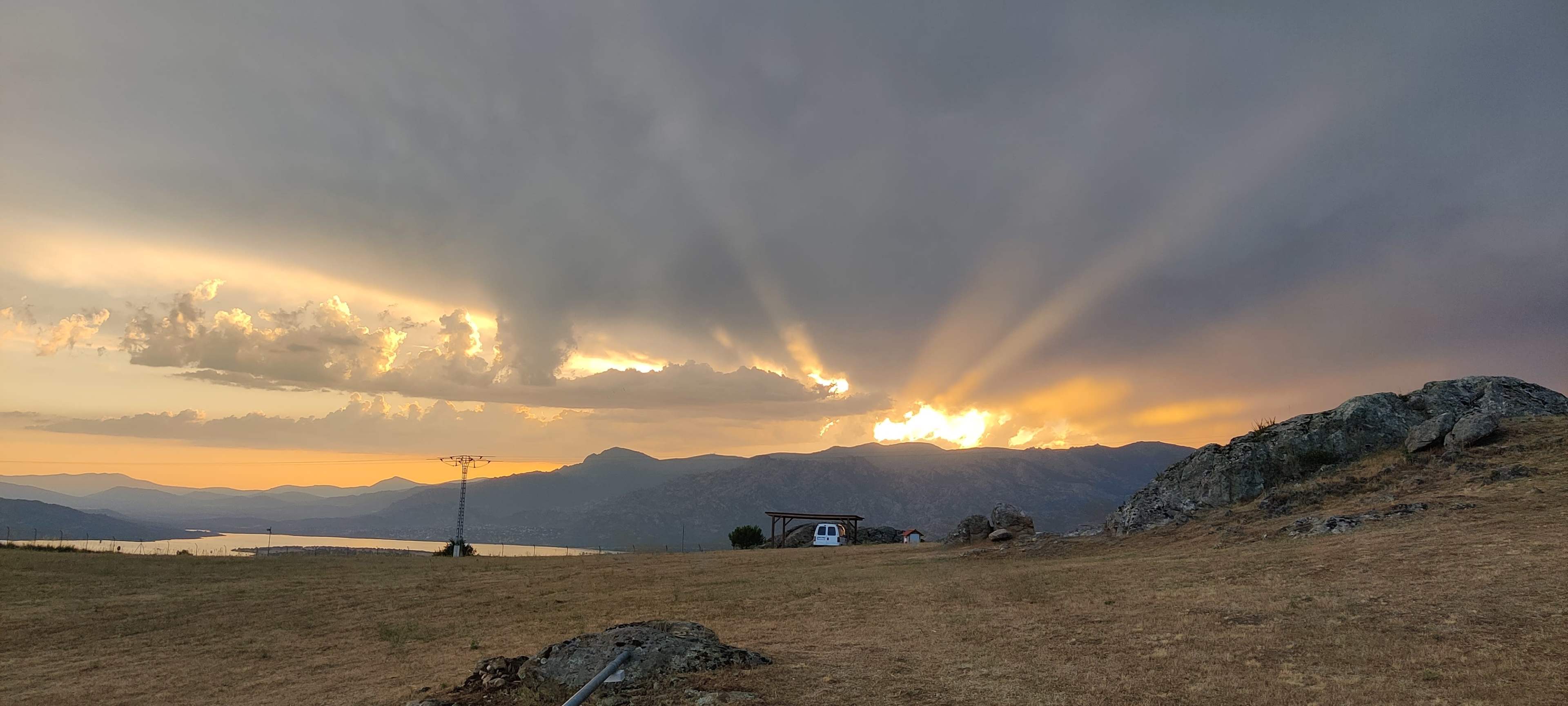 The image shows a sunset with rays of light emerging from behind a mountain range, while a small structure and a vehicle are visible on the foreground hillside.