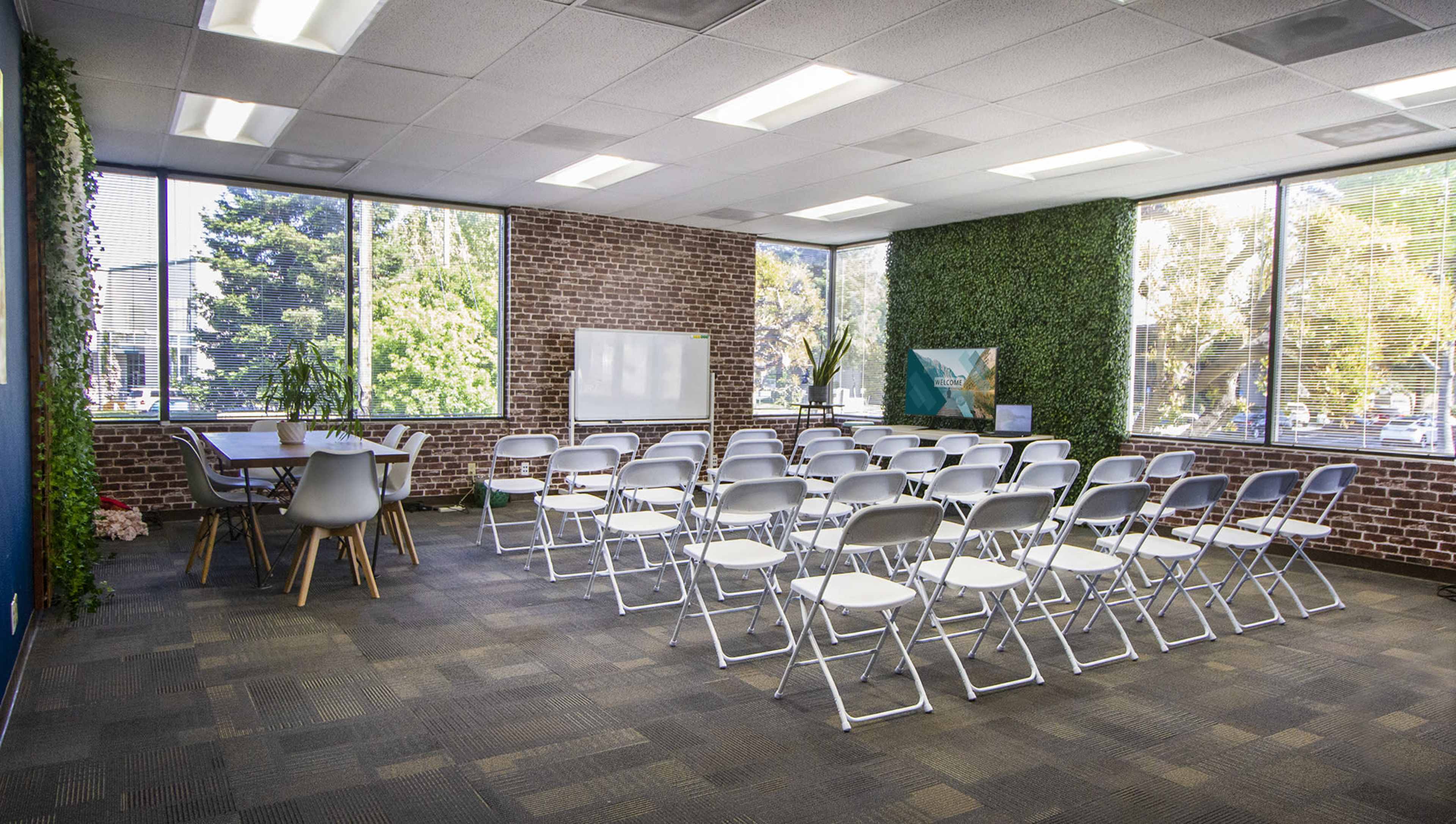 The image shows a well-lit meeting room with rows of white folding chairs arranged facing a whiteboard, surrounded by large windows and greenery on the walls.