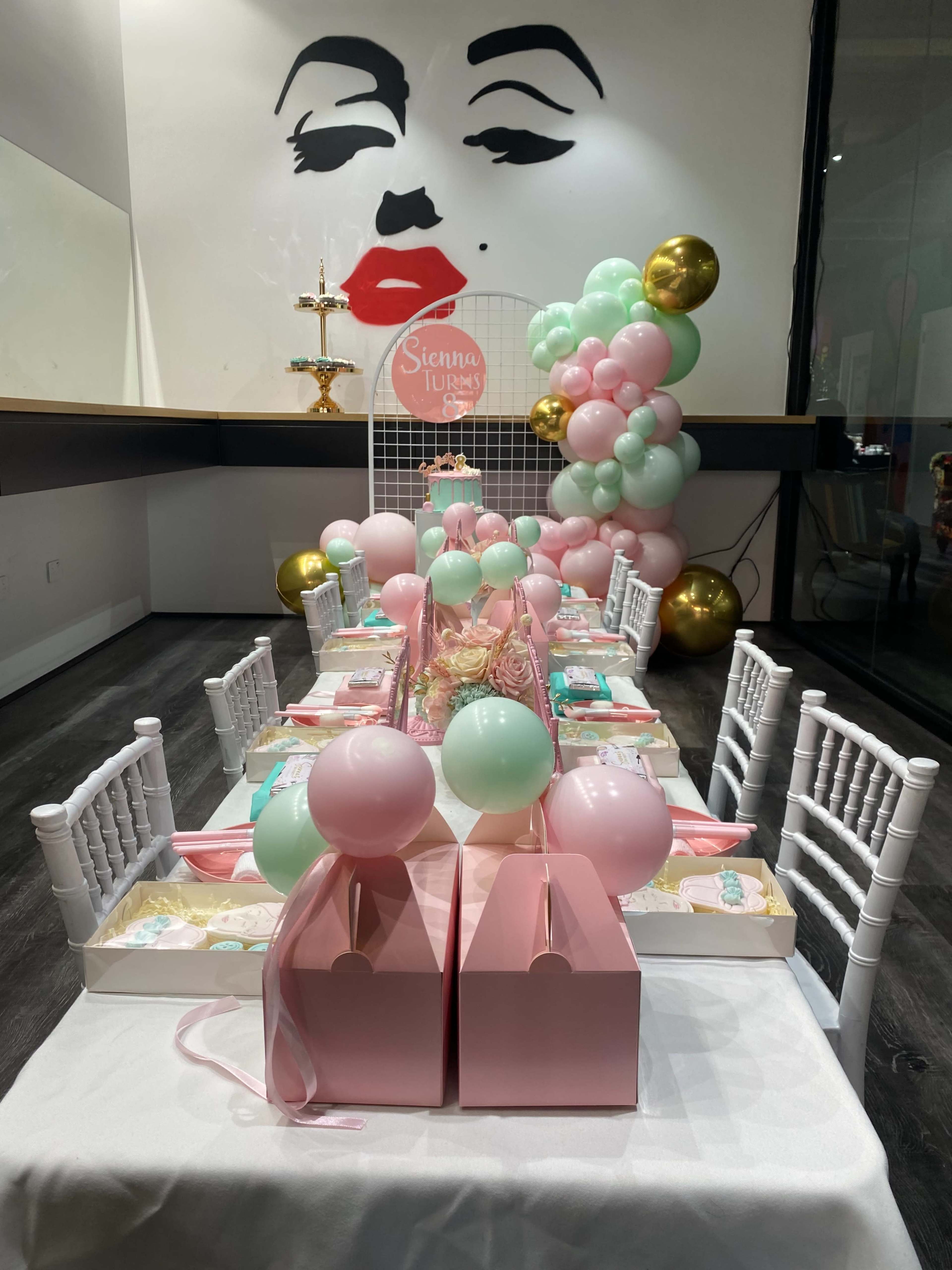 A long table is set for a party, adorned with pastel-colored decorations, gift boxes, and balloons, against a decorative wall featuring a large mural of a woman's face.