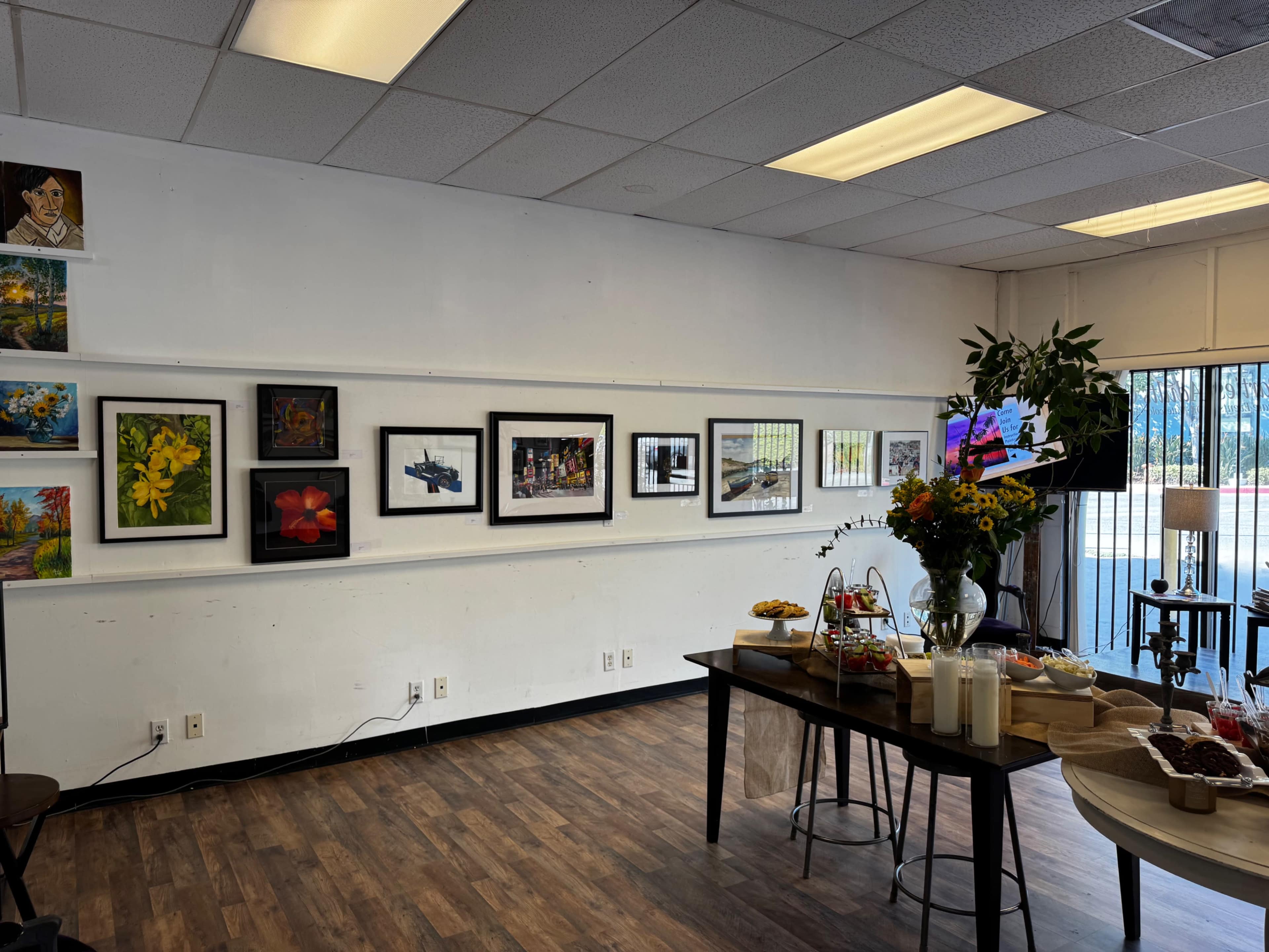 The image shows an art display on a wall featuring various framed paintings and photographs in a room with tables set up for a gathering.