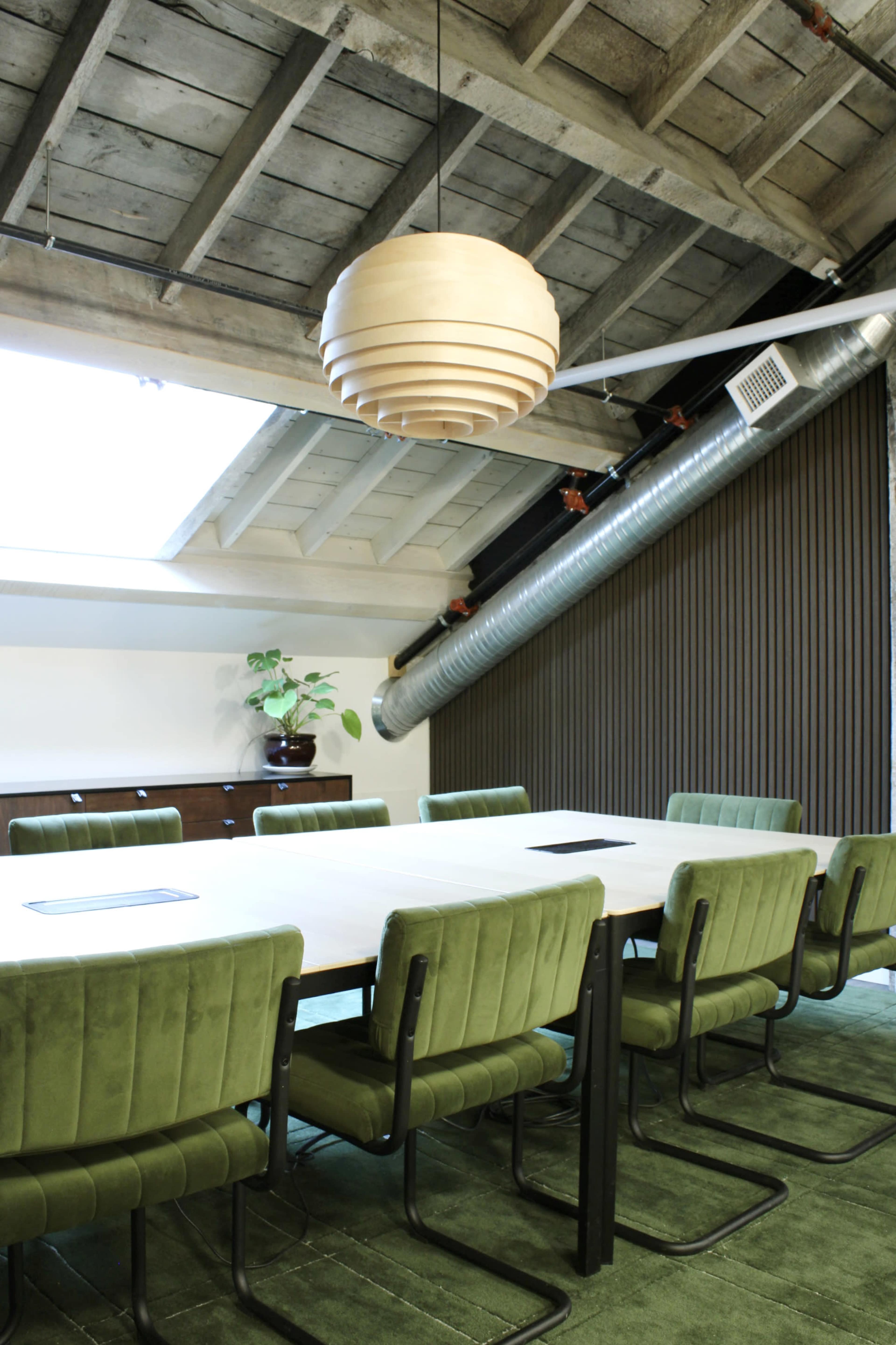 The image shows a modern conference room with a large table surrounded by green upholstered chairs and a skylight overhead, featuring exposed beams and an industrial-style metal duct.