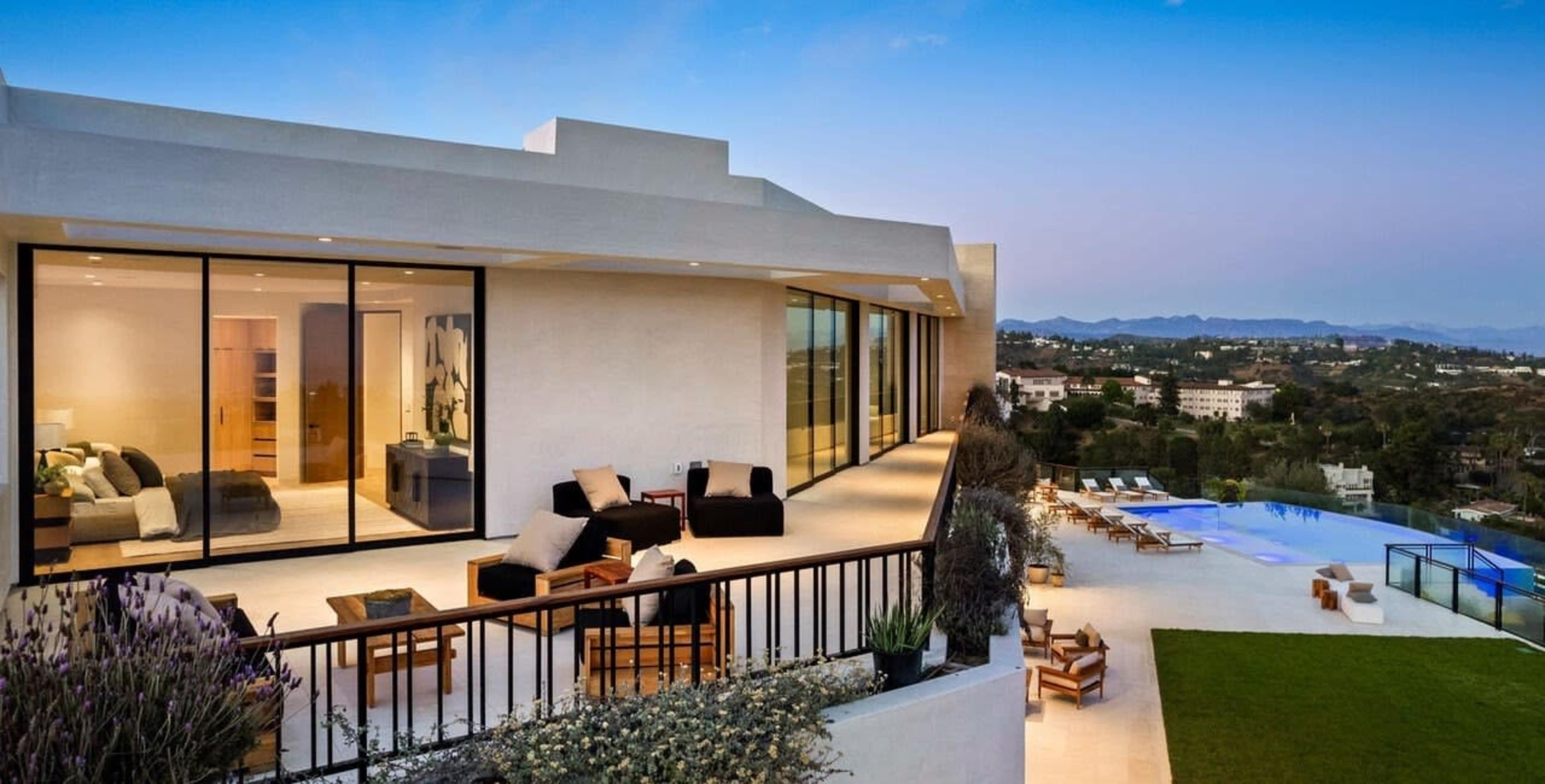 The image shows a modern house with large glass windows, featuring an outdoor seating area and a swimming pool overlooking a scenic landscape.