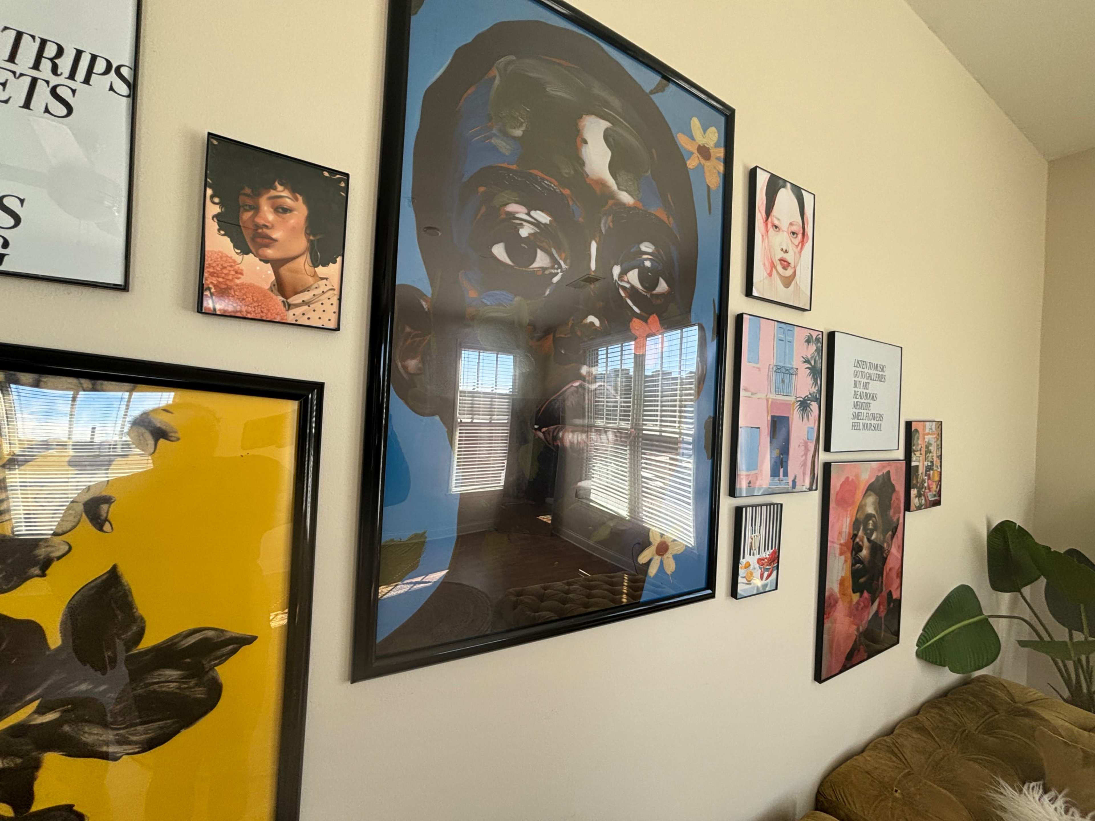 A wall adorned with various framed artworks, including portraits and abstract pieces, alongside a couch and large windows.