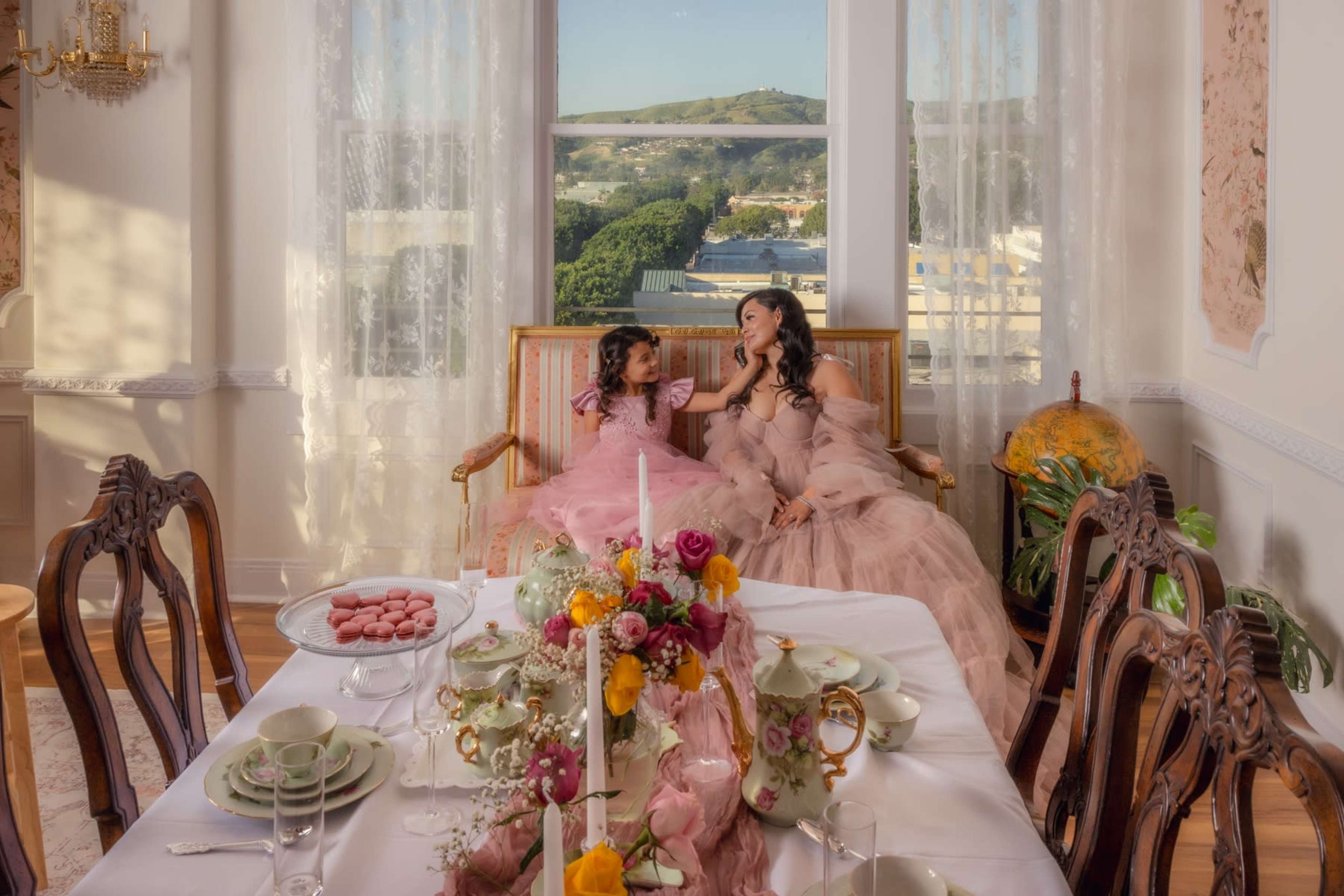 A woman and girl in pink dresses sit on a couch by a window, while a beautifully arranged dining table with floral decorations and tableware is set in front of them.