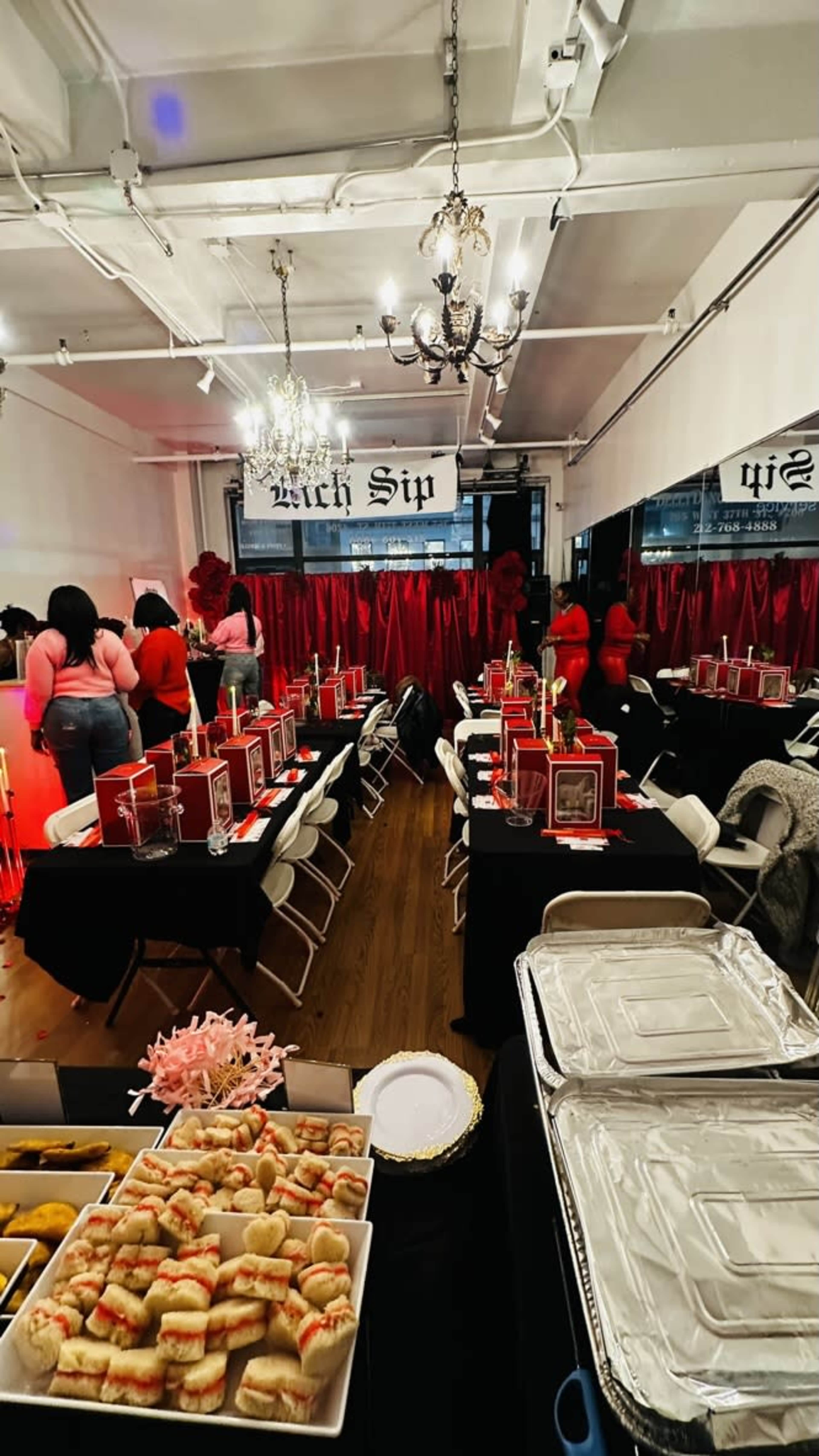 The image shows a decorated event space with tables set for a gathering, featuring food platters and attendees dressed in red.