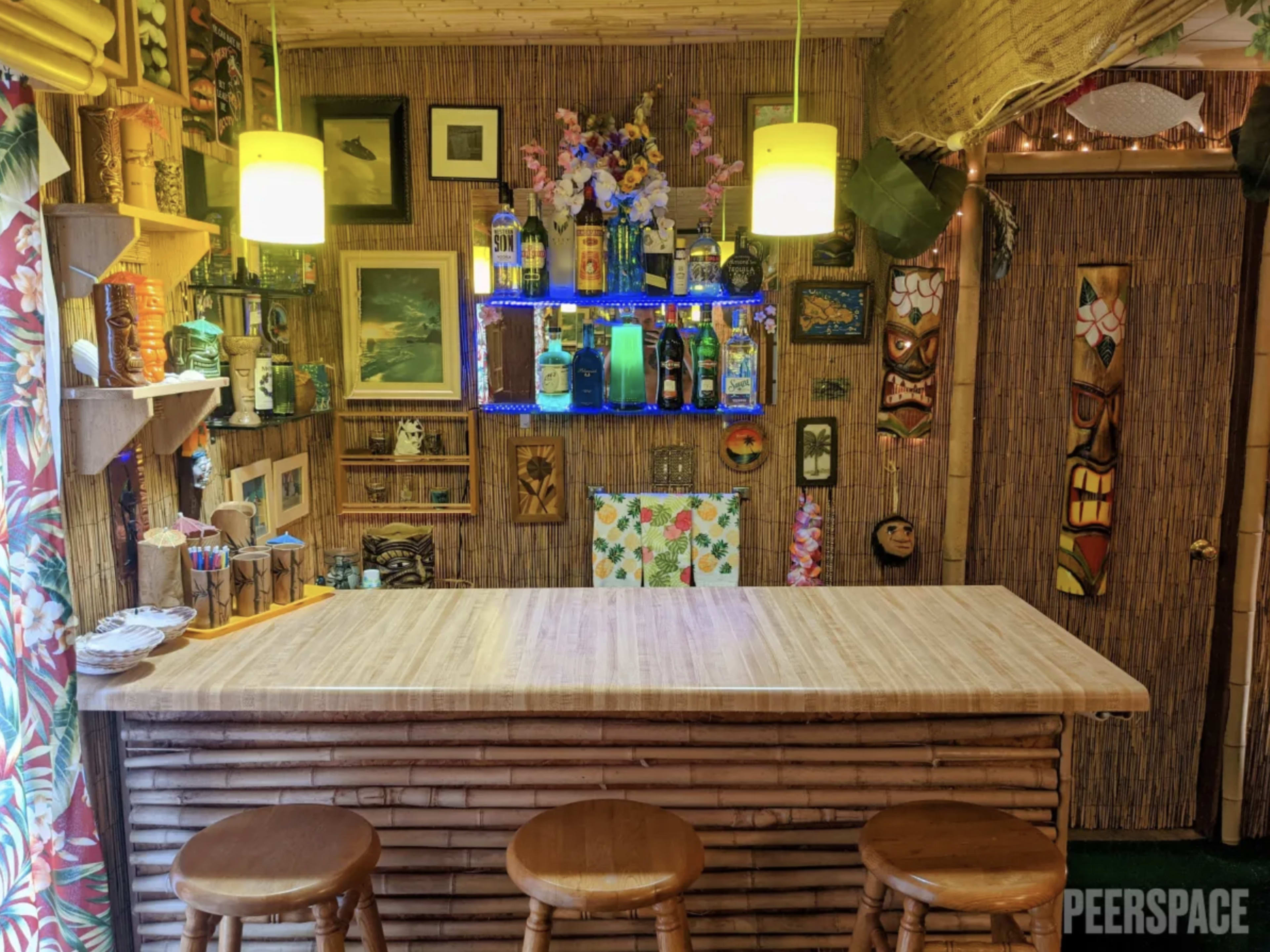 The image shows a tiki-themed bar with bamboo furnishings, colorful decorations, and a well-stocked liquor shelf illuminated by bright lights.