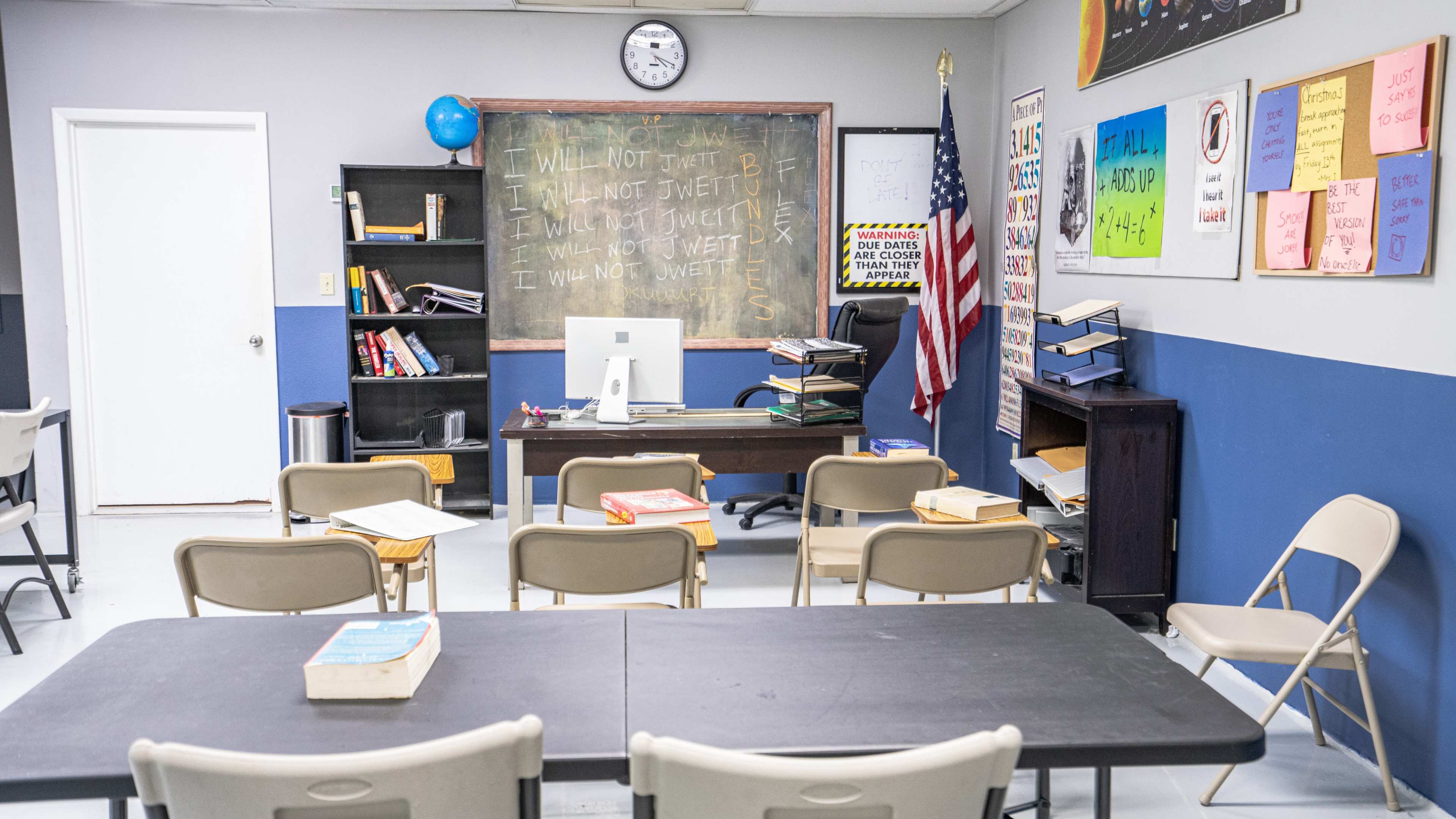 A classroom is set up with rows of chairs facing a desk, a chalkboard covered in writing, and an American flag on the wall.