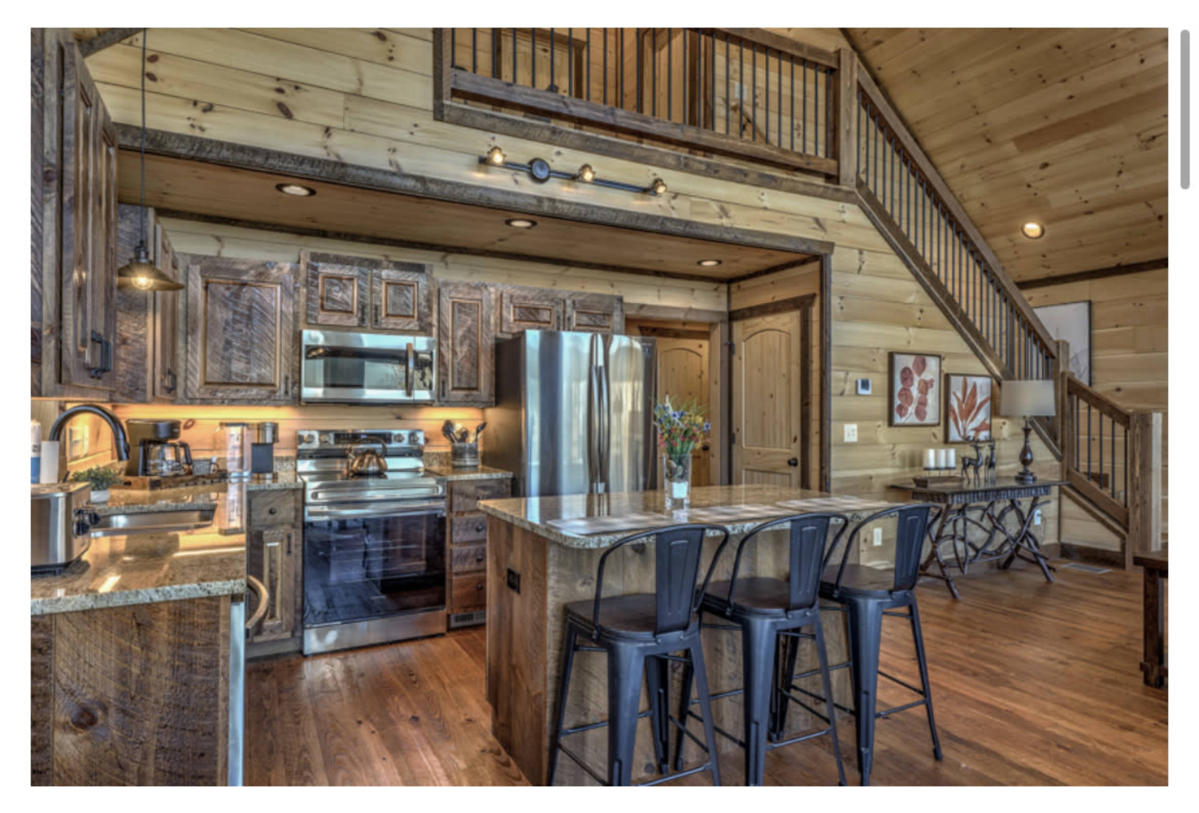 A wooden kitchen with rustic cabinetry, stainless steel appliances, and a central island with metal stools, leading to a staircase in a wooden interior.