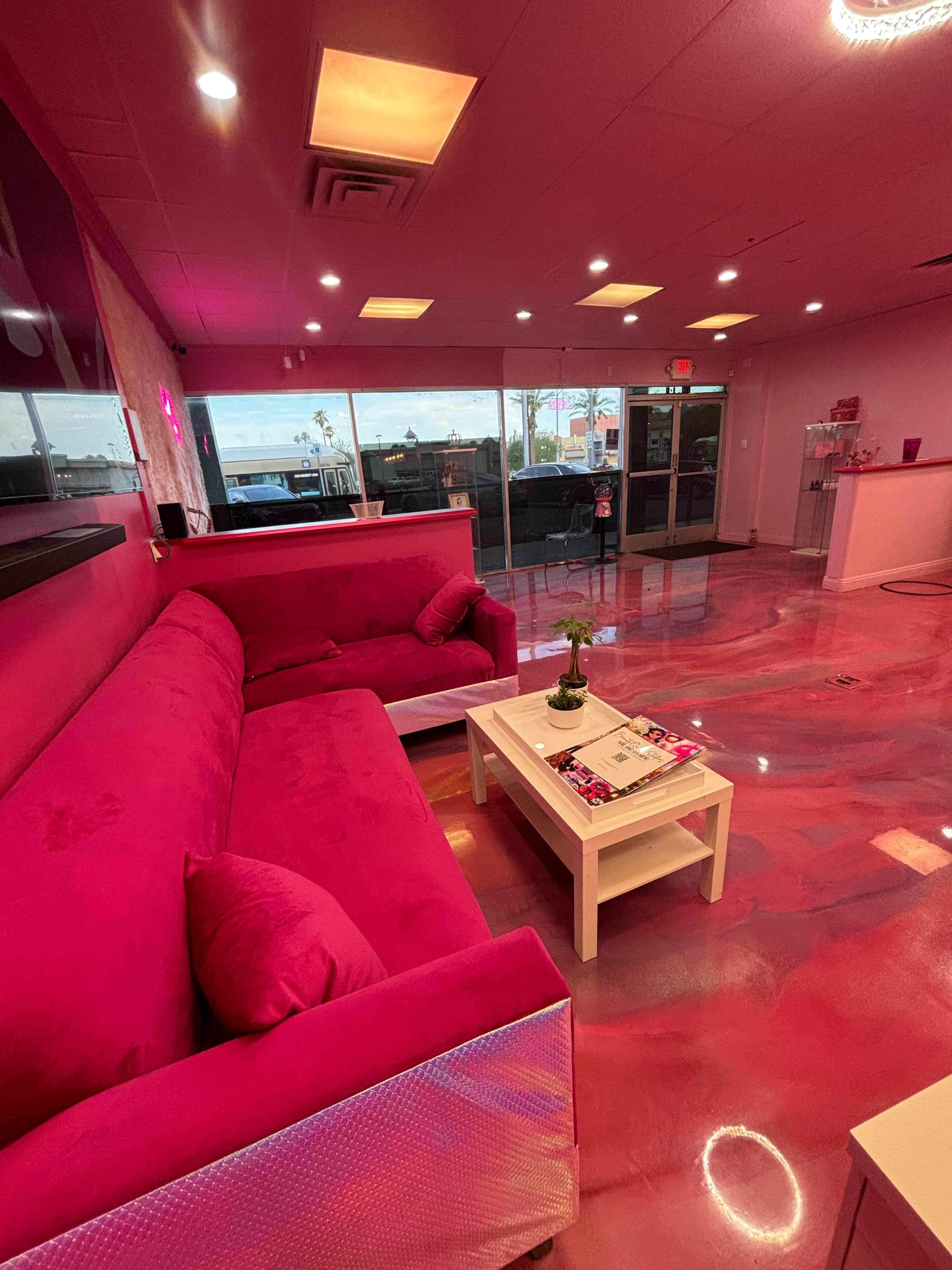 The image shows a vibrant pink lounge area with a pink couch and a small white coffee table, illuminated by soft lighting and large windows at the front.