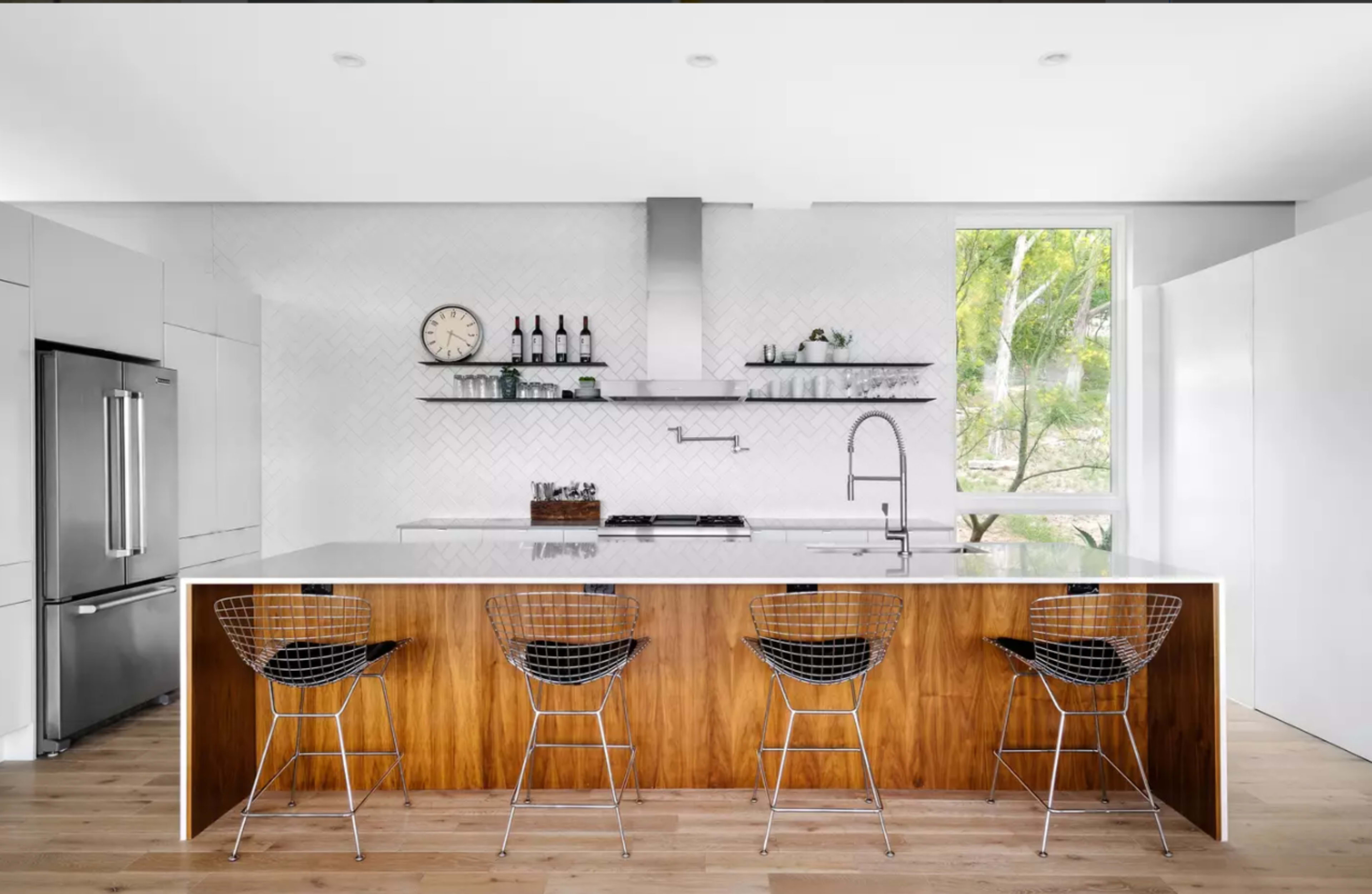 The image shows a modern kitchen featuring a large island with wooden accents, sleek stainless steel appliances, and wire bar stools.