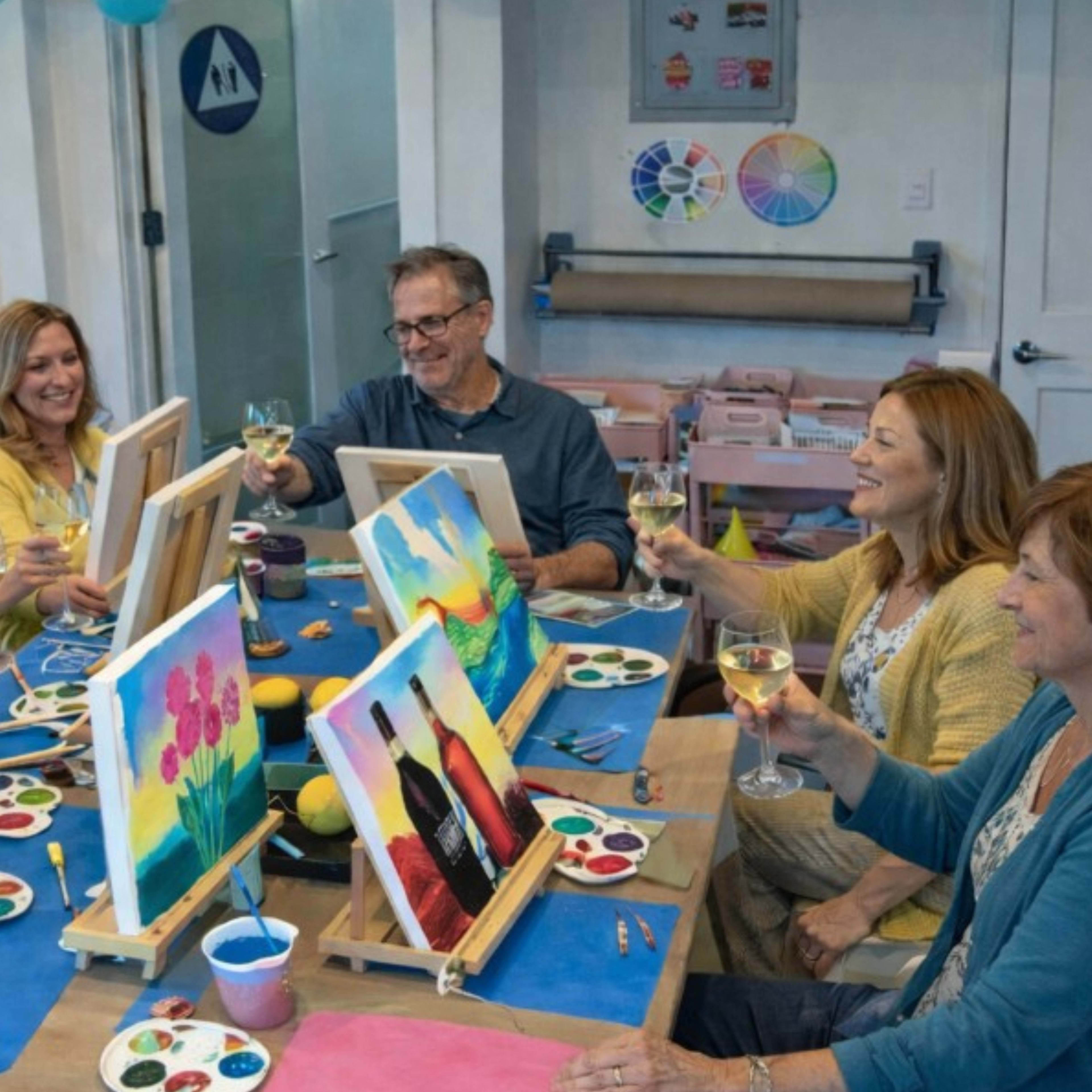 A group of five people is seated around a table, each holding a glass of wine and showcasing their colorful paintings on easels.