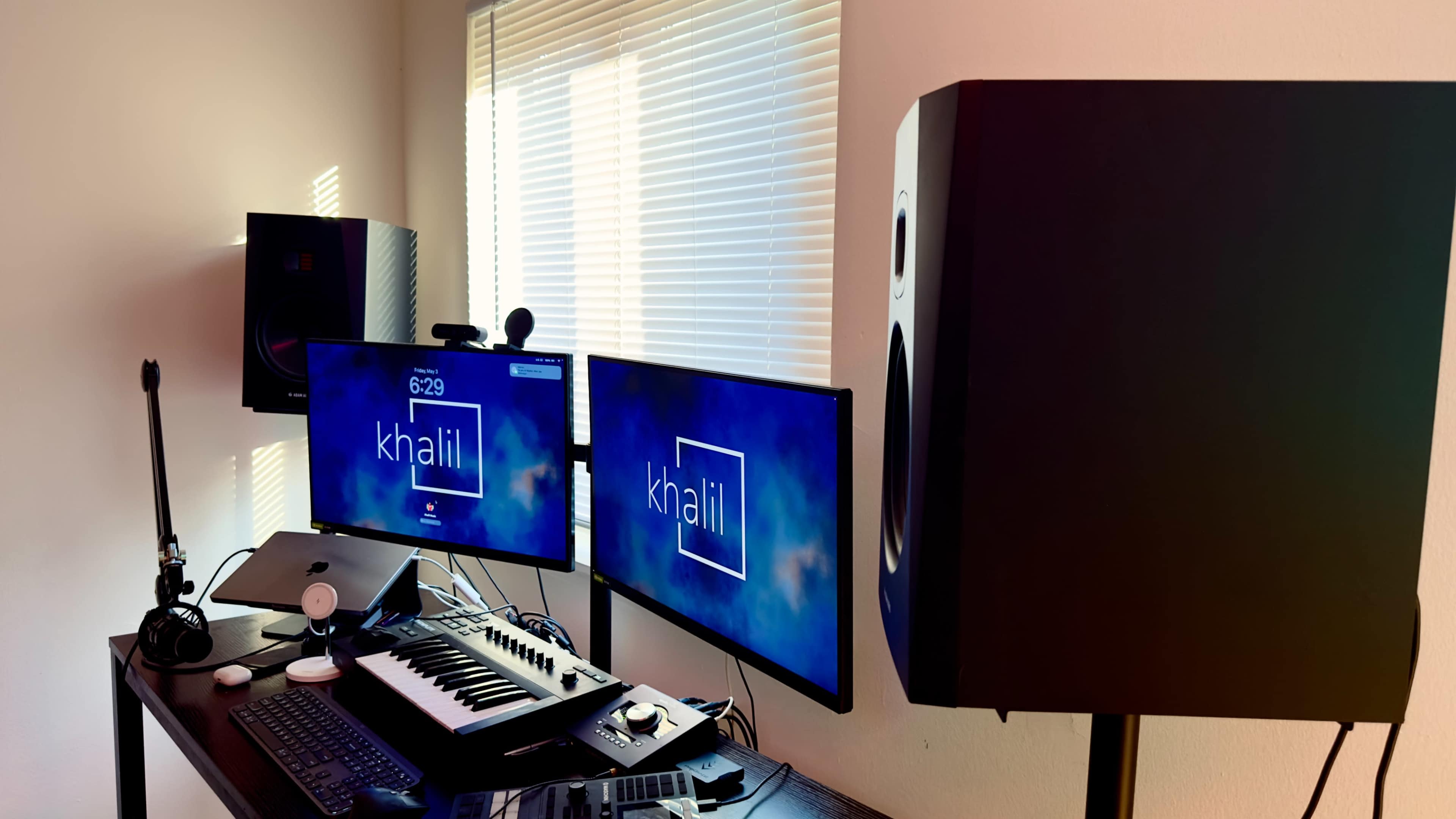 A modern music production workspace featuring two monitors, a keyboard, and studio speakers against a wall with window blinds.
