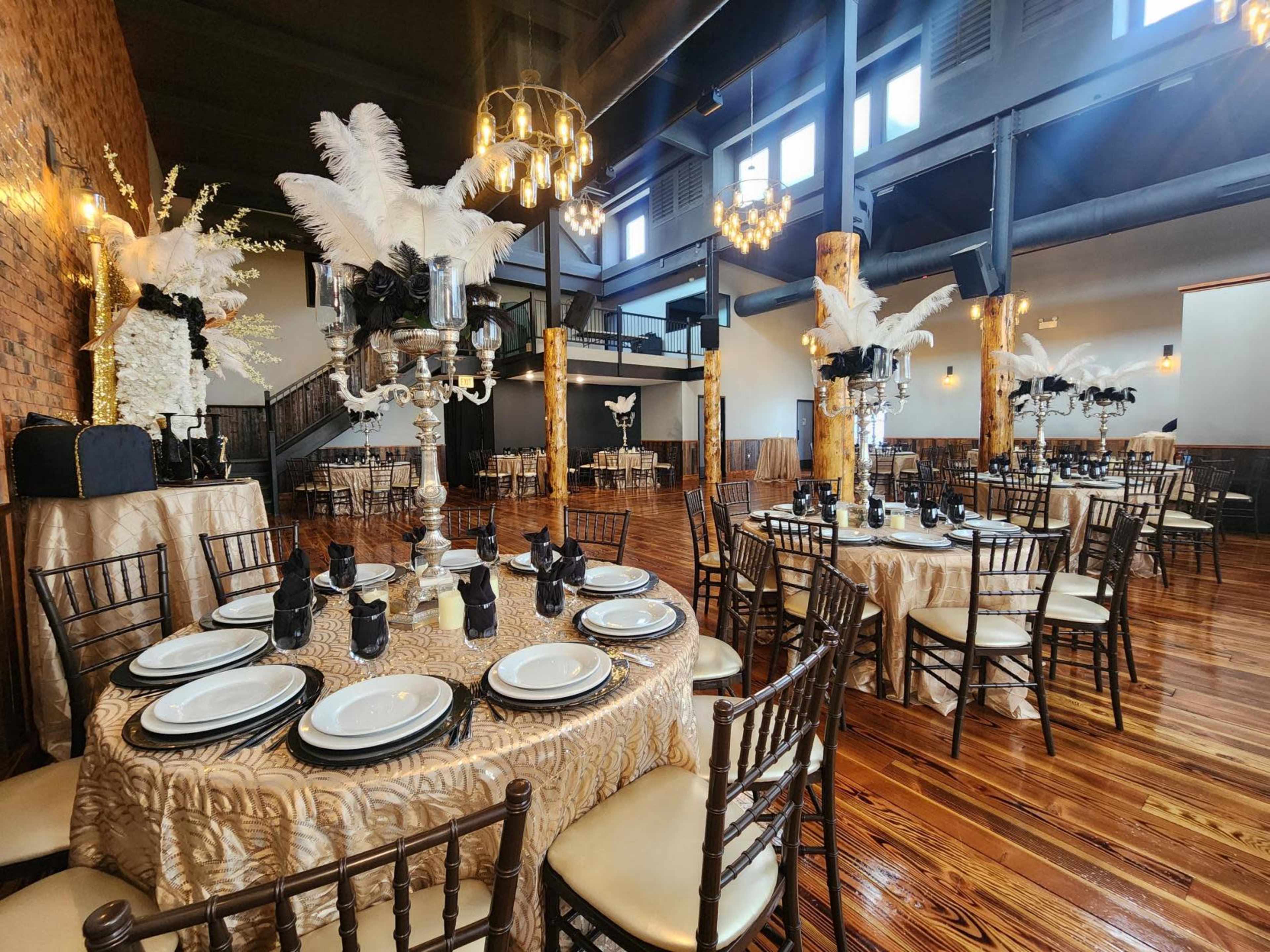 An elegantly arranged banquet hall with several round tables set with plates and black napkins, surrounded by tall decorative centerpieces and ambient lighting.