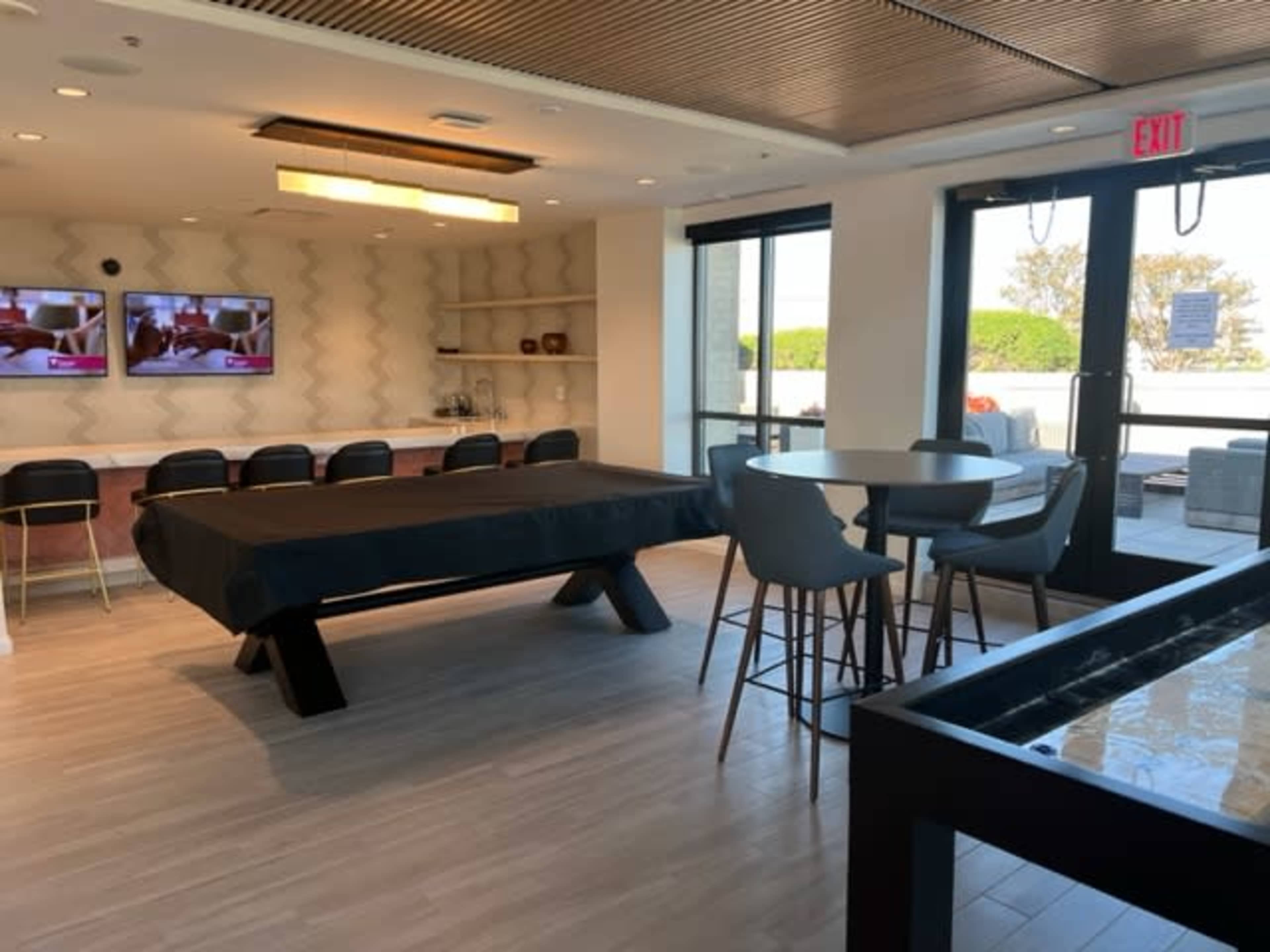 A modern indoor lounge area featuring a billiards table, two television screens mounted on the wall, and a seating arrangement with high tables and chairs.